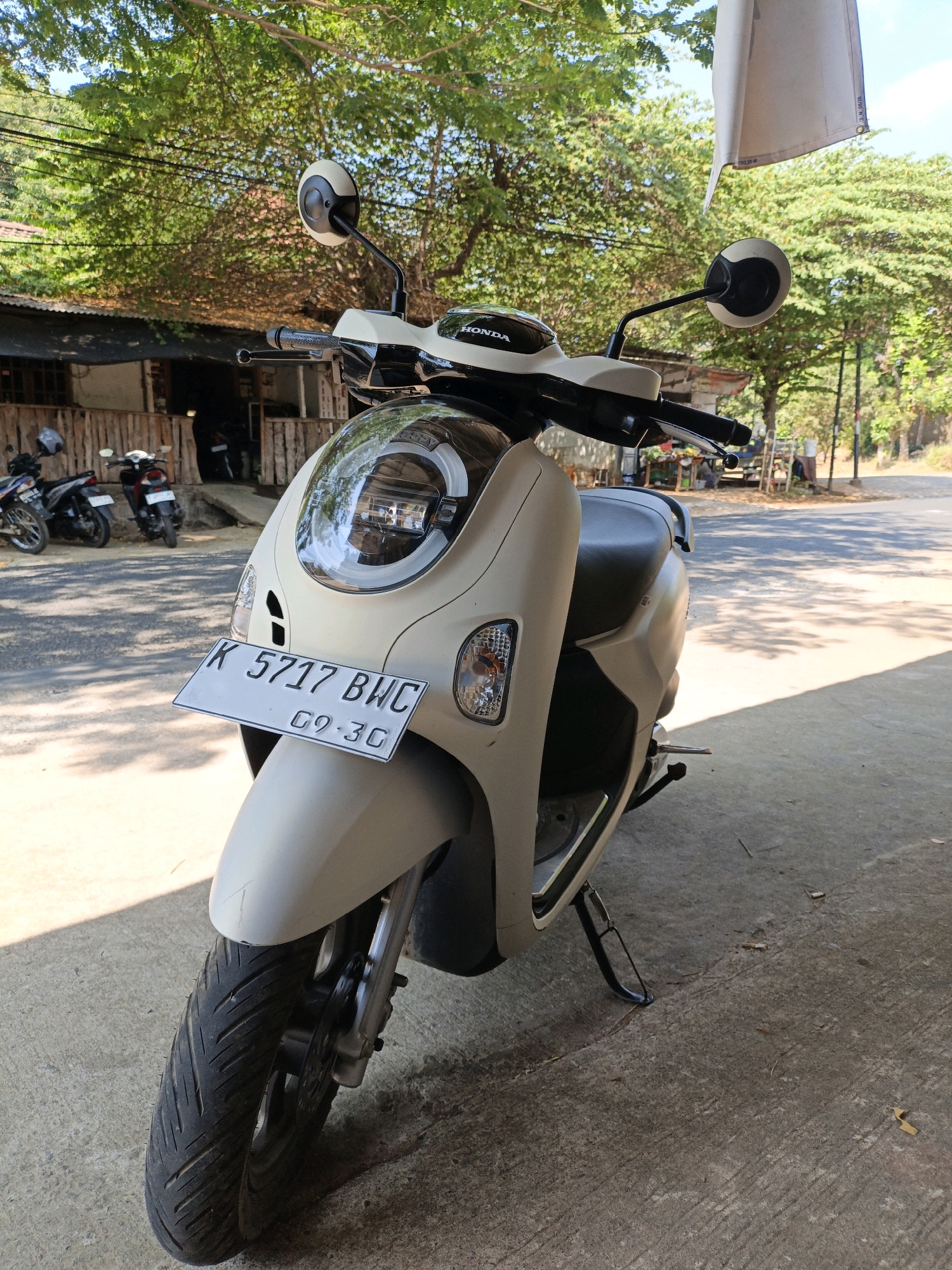 A cream-colored Honda scooter is parked on a concrete surface under the shade of trees. The scooter faces the viewer, with its front wheel clearly visible. A license plate reading "K 5717 BWC 09.30" is attached to the front. In the background, there are other parked motorcycles, a building with a corrugated metal roof, and a road. The environment appears to be an outdoor area, possibly a roadside establishment in Bandengan, Indonesia. Sunlight filters through the leaves of the trees, creating dappled shadows on the ground.
