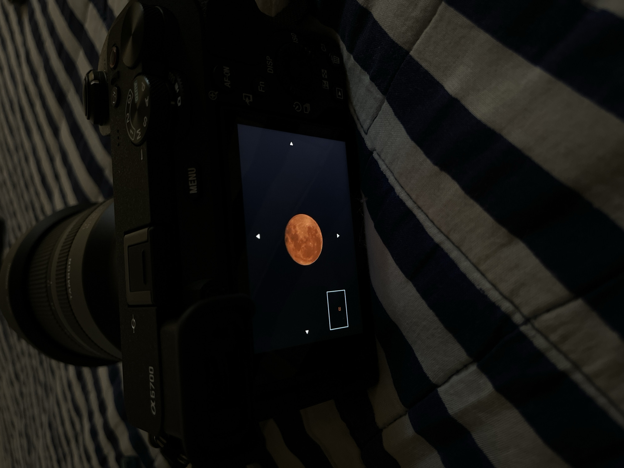 Here is a forensic description of the image:
The image shows a Sony a6700 camera placed on a surface with blue and white stripes, possibly fabric. The camera's LCD screen displays a photograph of a full, reddish moon against a black background. The screen also shows interface elements like directional arrows and a focus box. The camera is equipped with a Tamron lens. The scene is dimly lit, highlighting the camera screen. The location of San Bernardino Tlaxcalancingo, Mexico is not evident within the picture's content.