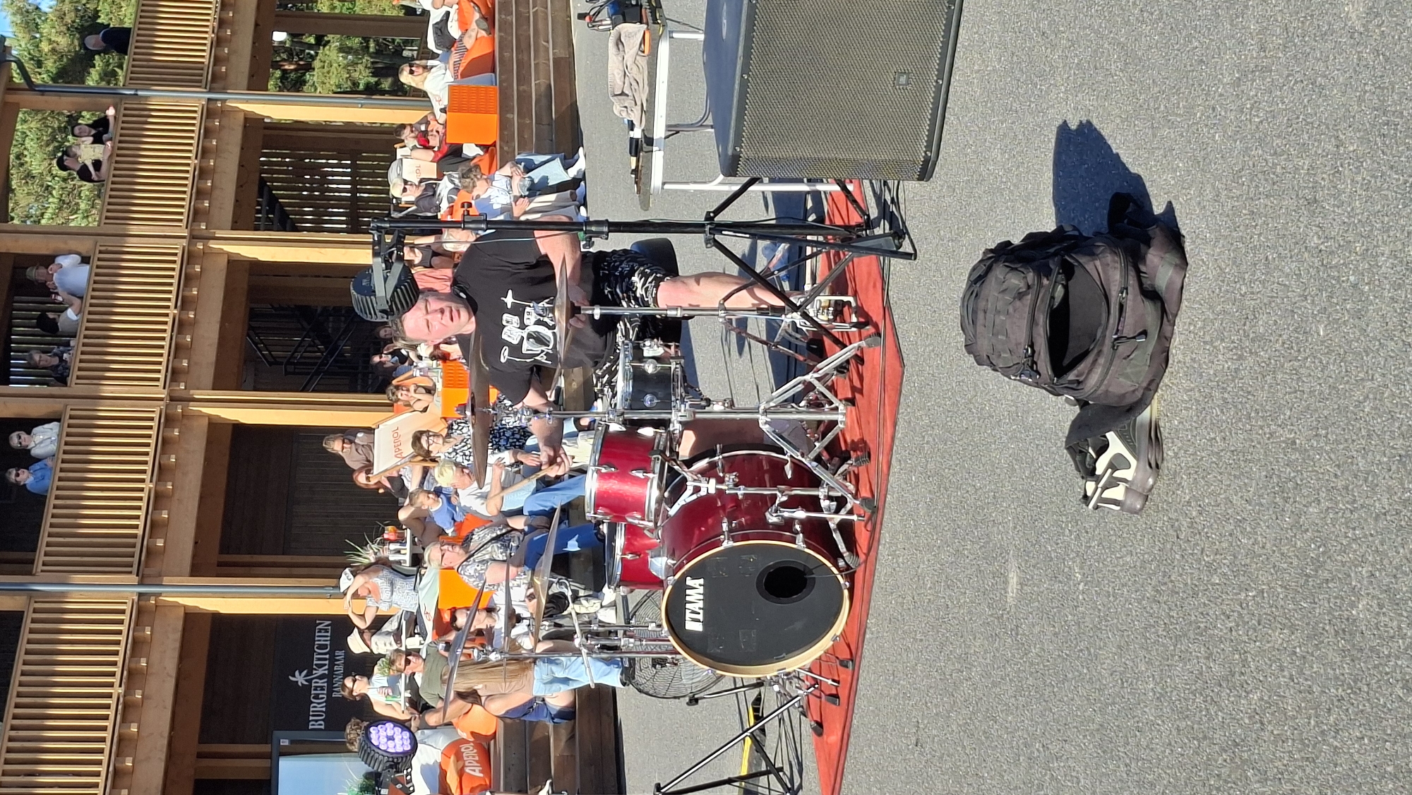 The image shows an outdoor scene with a drum set in the foreground, a drummer playing, and an audience in the background. The drummer, a man with short hair, is wearing a black t-shirt with a graphic print and patterned shorts. He is seated behind a red and black drum kit, holding drumsticks. A fan is positioned to the left of the drum set. In the background, a multi-level wooden structure with balconies is visible, occupied by people watching the performance. Some individuals are seated on orange cushions. The setting appears to be a public space or venue, possibly a restaurant or entertainment complex, given the signage for "BURGER KITCHEN BANNABAAR" and "APEROL" on a screen and advertising banners. A backpack and a pair of shoes are placed on the asphalt in the lower right portion of the frame. The overall environment suggests a warm, sunny day with a casual, lively atmosphere. The scene is consistent with an outdoor musical performance in Tallinn, Estonia, likely during the summer season.