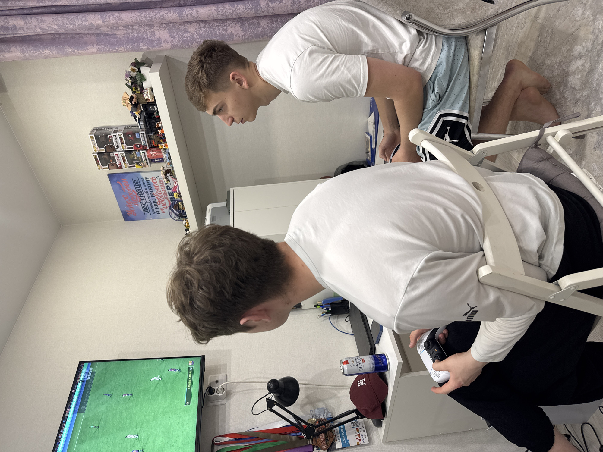 Two young men are engaged in a video game in an indoor room, likely in Kazan, Russia. One man is seated, actively playing with a white PlayStation controller, while the other stands beside him, intently watching the game on the television screen. The game displayed is a football (soccer) simulation, with text "LAMINE YAMAL 19" visible, indicating a player's name and number, and "4 СМЕНА ИСПОЛНИТЕЛЯ" (4th PLAYER CHANGE).
The setting appears to be a cozy living space or bedroom, characterized by light-colored walls and a light purple curtain. A white shelf above the gaming area holds various objects, including several Funko Pop figures, with "SHAQUILLE O'NEAL" and "DEADPOOL" identifiable. Below the shelf, a poster or sign features text in Russian: "Успех порожден лестнице приставленной к стене И ЕЩЕ НИКОМУ не удавалось ВЗОБРАТЬСЯ ВВЕРХ" (Success is born from a ladder leaning against a wall AND NO ONE has ever managed TO CLIMB UP).
On the desk or table next to the seated player, a small black lamp, a red baseball cap, and a blue and silver can labeled "KRONE BLANC" are visible. A small sign or card near some medals reads "КАЗБА" (KAZBA) and "СПОРТСМЕН" (SPORTSMAN). Both individuals are focused on the game, suggesting an immersive experience. The image is taken indoors, with no clear indication of the time of day or weather.