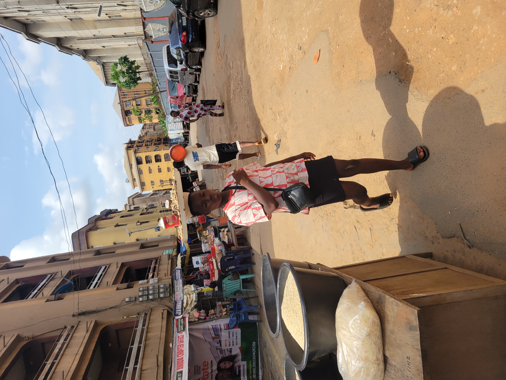 In Onitsha, Nigeria, a young person walks towards the camera on a dusty, uneven street. They are wearing a red and white patterned short-sleeved shirt, a black short, and black slip-on sandals. A black shoulder bag is slung across their chest. To their left, large black basins filled with grain are visible on a wooden counter. In the background, a street market is in full swing with various stalls and people. Buildings line the street, some appearing to be multi-story residential or commercial structures. Cars are parked along the roadside, and overhead wires crisscross the sky. The scene is bustling with activity, suggesting a typical day in a Nigerian city.