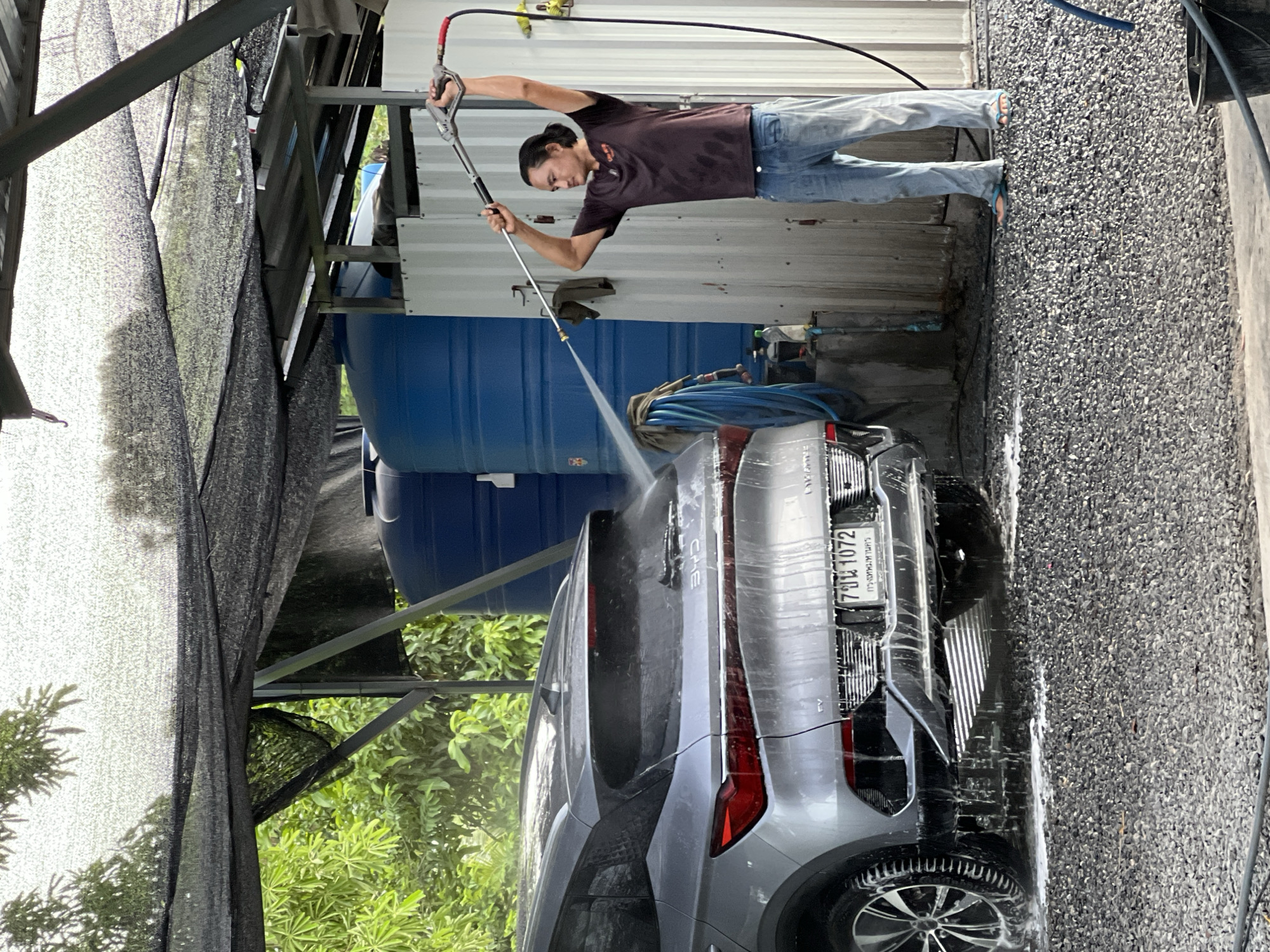 A man is power washing the back of a gray SUV at a car wash in Ban Bang Rak, Thailand. The man, with dark hair pulled back in a bun, wears a dark t-shirt and light blue jeans, and stands to the right of the vehicle, holding the wand of a power washer above his head with both hands. Water is spraying from the wand onto the car's rear. The SUV is parked on a gravel surface, with a lot of foam and water running down its sides and onto the ground. Behind the car are large blue water tanks and a corrugated metal wall. Green foliage is visible in the background, partially obscured by a black mesh shade structure covering the car wash area. A license plate on the rear of the car reads "7 ขน 1072".