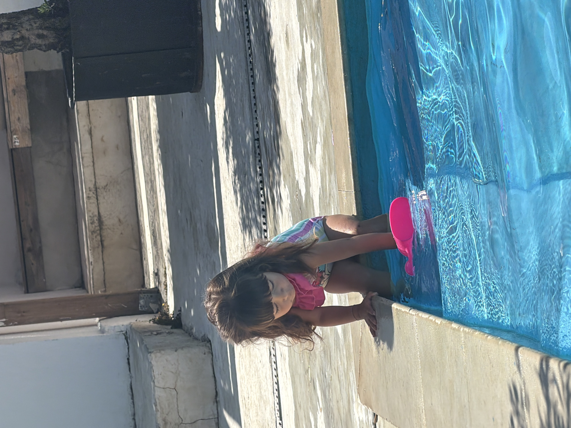 A young girl with long brown hair is leaning over the edge of a swimming pool, reaching into the blue water. She is wearing a swimsuit with a pink ruffled top and a striped bottom. In her right hand, she holds a bright pink watering can. Her gaze is directed downwards into the pool. The scene appears to be outdoors, with sunlight casting shadows from what looks like trees or foliage onto the pool deck. The pool deck is made of light-colored concrete, and there are some stone steps or a raised platform to the left of the girl. The water in the pool is clear and reflects the sunlight, creating shimmering patterns. The overall impression is a warm, sunny day with the girl enjoying playtime by the pool. The image appears to be taken in Burgas, Bulgaria. No visible text is present in the image.