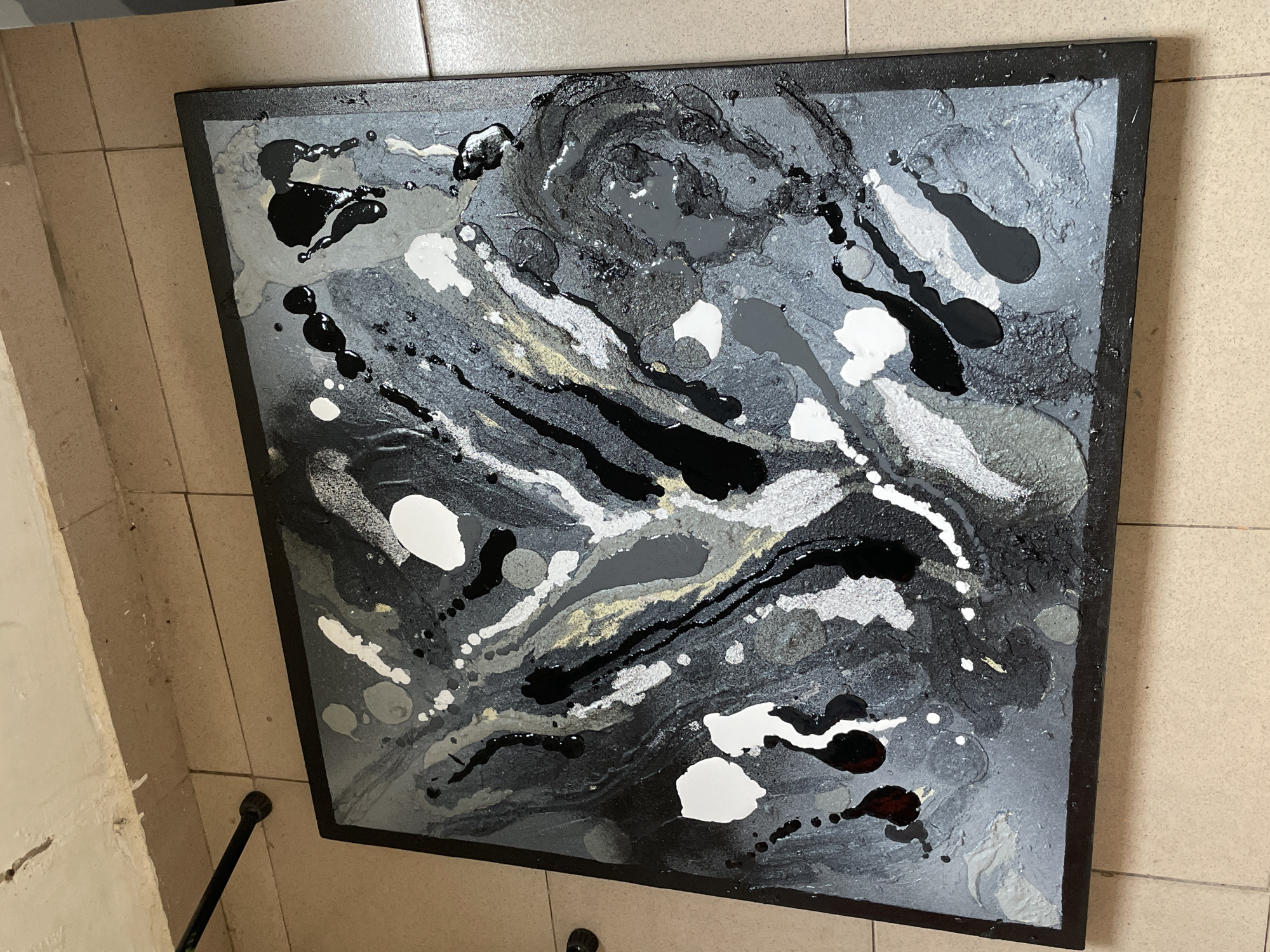 The image shows a square abstract painting leaning against a wall with light beige tile flooring. The painting has a black frame and is primarily black, white, grey and silver. The patterns consist of swirling lines, blobs, and splatters. The floor is tiled, and the wall is a light beige colour. A dark metal pole is visible in the bottom left corner of the image. It is unclear where this scene occurs in the city of Ado, Nigeria. No people or actions are depicted.