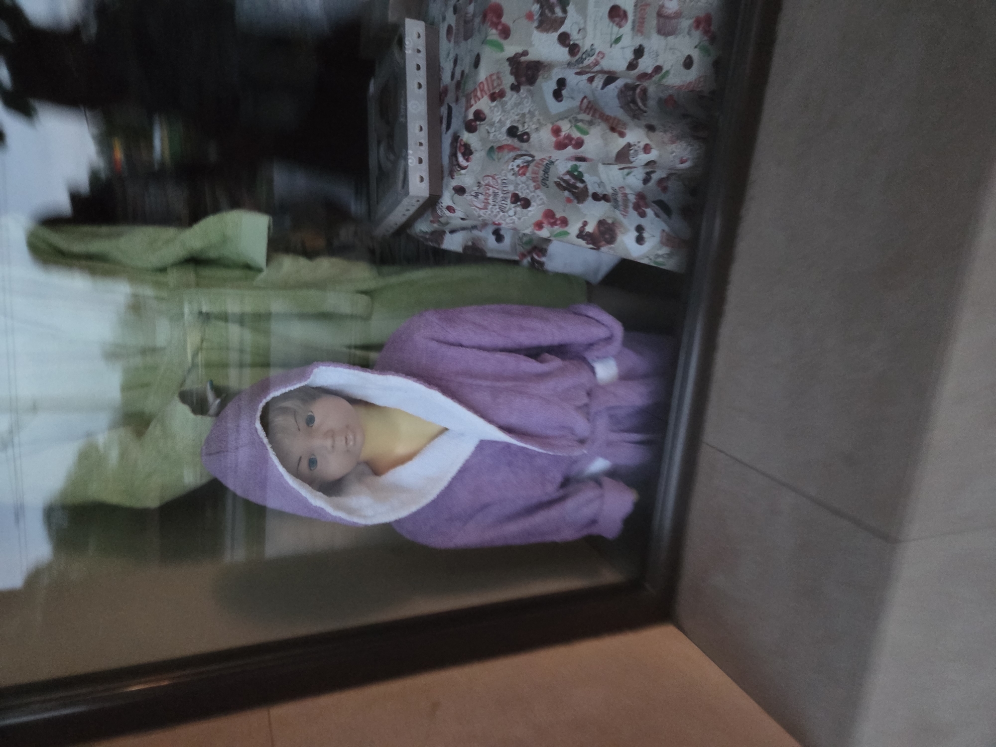 The image displays an indoor scene, likely a shop window or a display case, captured through a reflective glass surface. This causes notable reflections and some blurring, particularly in the background.
The central subject is a baby doll, depicted lying on its side (the image itself is rotated 90 degrees counter-clockwise). The doll has blue eyes and light-toned skin, and is dressed in a soft, hooded purple robe with white trim.
Behind the doll and to its left, a folded green textile, possibly a towel or blanket, is visible. In the upper portion of the image, behind the green textile and extending to the right, is a patterned fabric, likely a tablecloth. This fabric features a design of red and dark cherries, interspersed with cupcake illustrations. The word "CHERRIES" is clearly visible on this fabric, along with other text that appears to relate to desserts or sweets. Partially obscured behind the patterned fabric is a light-colored, possibly wooden or cardboard, box with small, circular holes along one edge.
The foreground of the image is dominated by a dark, thick frame, which appears to be part of the display structure or window. To the right and bottom of this frame, a plain, light brown or tan wall or floor is visible. The background behind the displayed items is largely indistinct and dark, further obscured by the reflections on the glass. The scene is static, suggesting items arranged for sale or exhibition. There are no human subjects, and no specific time of day or weather conditions can be discerned from the indoor lighting.