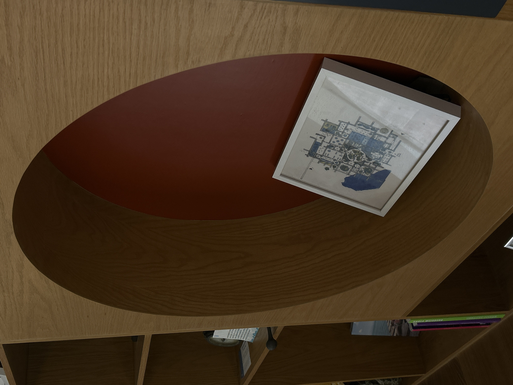 The image shows an overhead view of a wooden coffee table with an oval-shaped depression featuring a red inner surface. Resting partially on the red surface and partially on the surrounding wood is a framed picture with a white border, depicting what appears to be a schematic drawing with blue details. The table is part of a shelving unit, with books and other items visible on the shelves below. The location in Mazatlán, Mexico, is not discernible from the image alone.