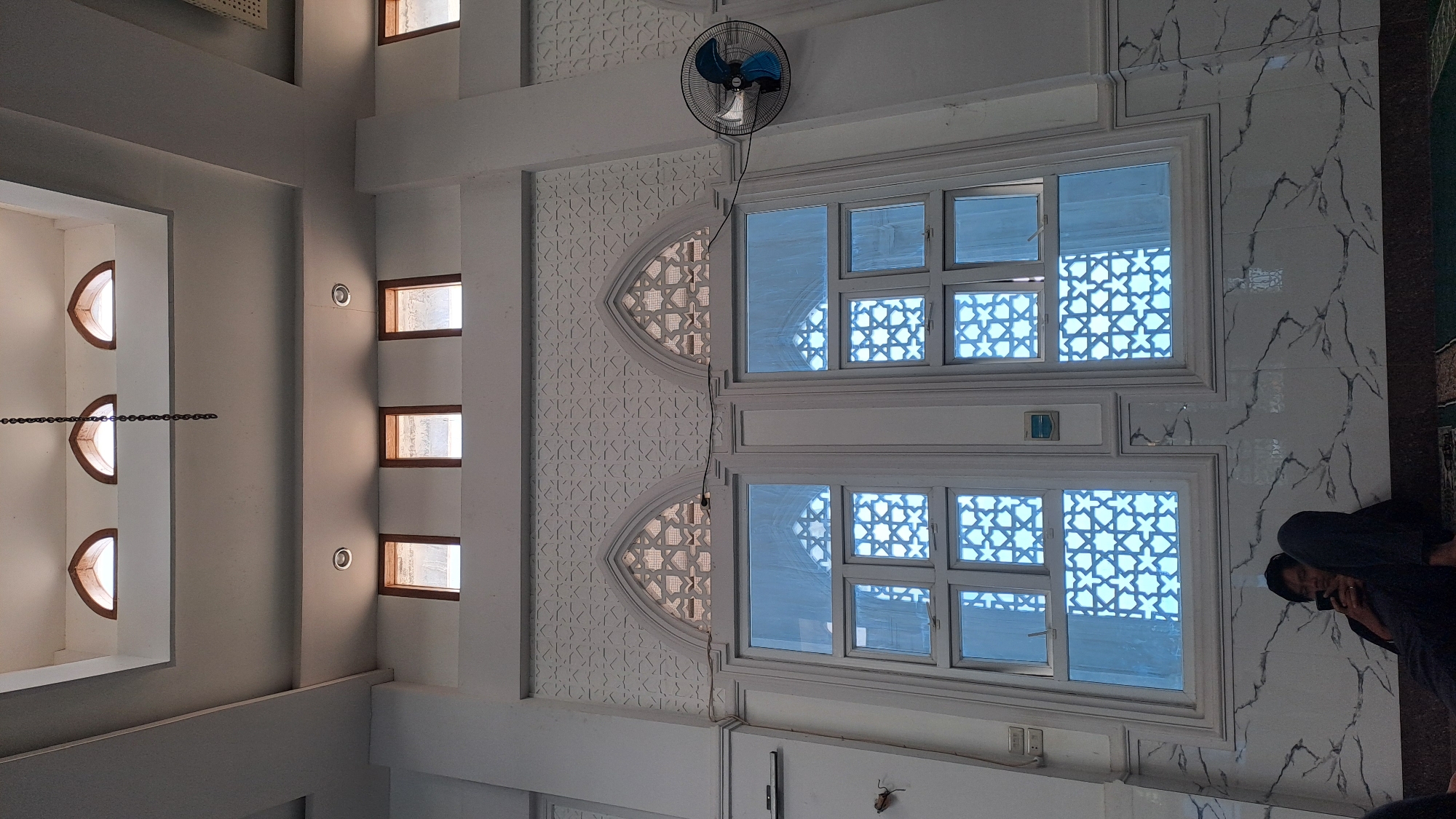 The image depicts the interior of a building, likely a mosque, in Kalalengkio, Indonesia. The scene is characterized by intricate architectural details. The ceiling features a geometric pattern with arched windows, and a chain hangs from the center. Below, a large window with decorative geometric patterns dominates the frame. The lower section of the wall is covered in marble with a veined pattern. A person, dressed in dark clothing, is seated on the floor, looking at their phone. A black wall-mounted fan is visible on the right side of the frame. The overall lighting suggests an indoor environment.