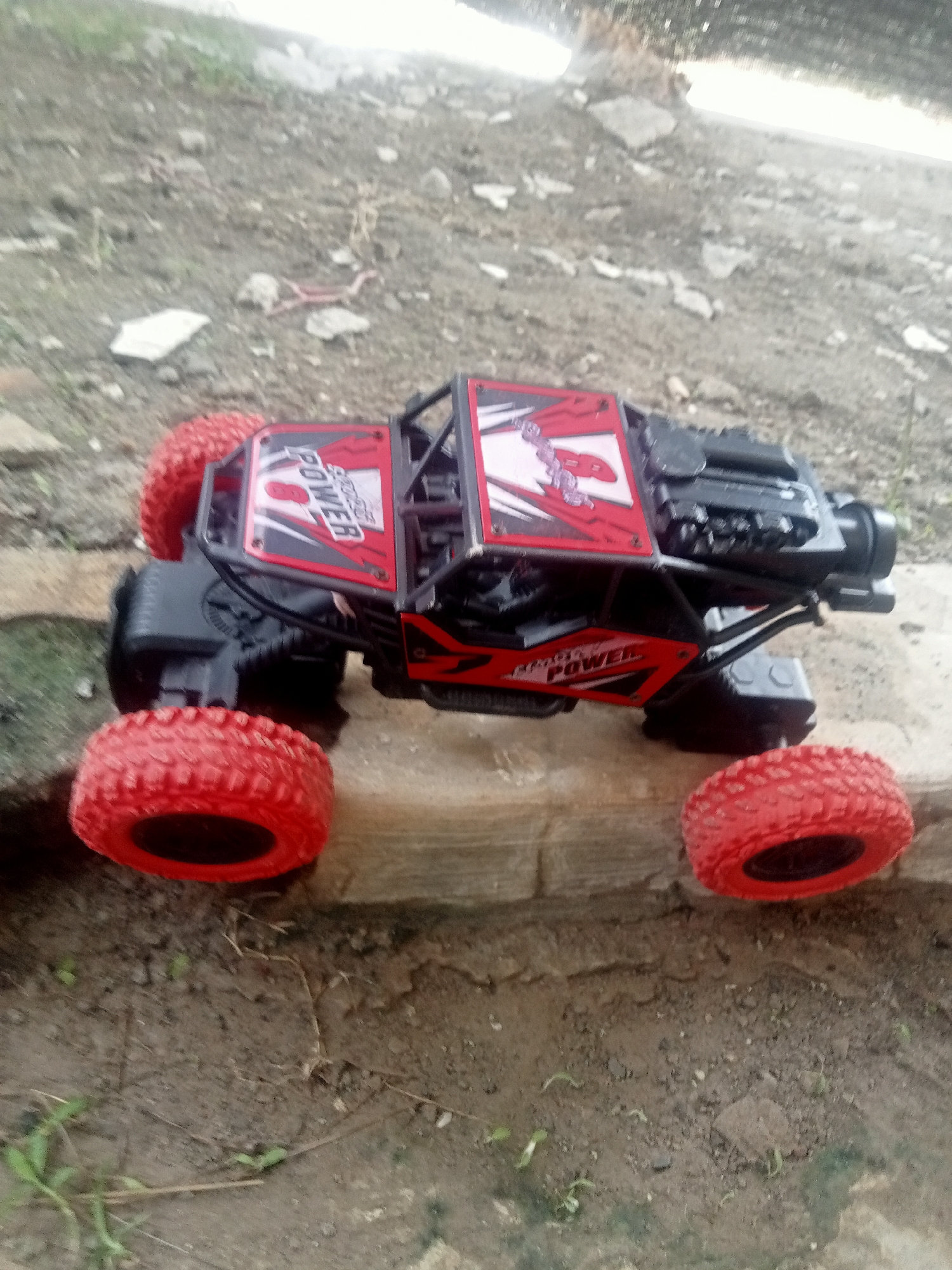 A red and black remote-controlled toy car, labeled with "POWER," rests on a light-colored, raised surface. The car has large, red, knobby tires. The background appears to be an outdoor, earthy environment with scattered rocks and sparse vegetation. The scene is likely located in Kutosari, Indonesia, as indicated by the provided context.