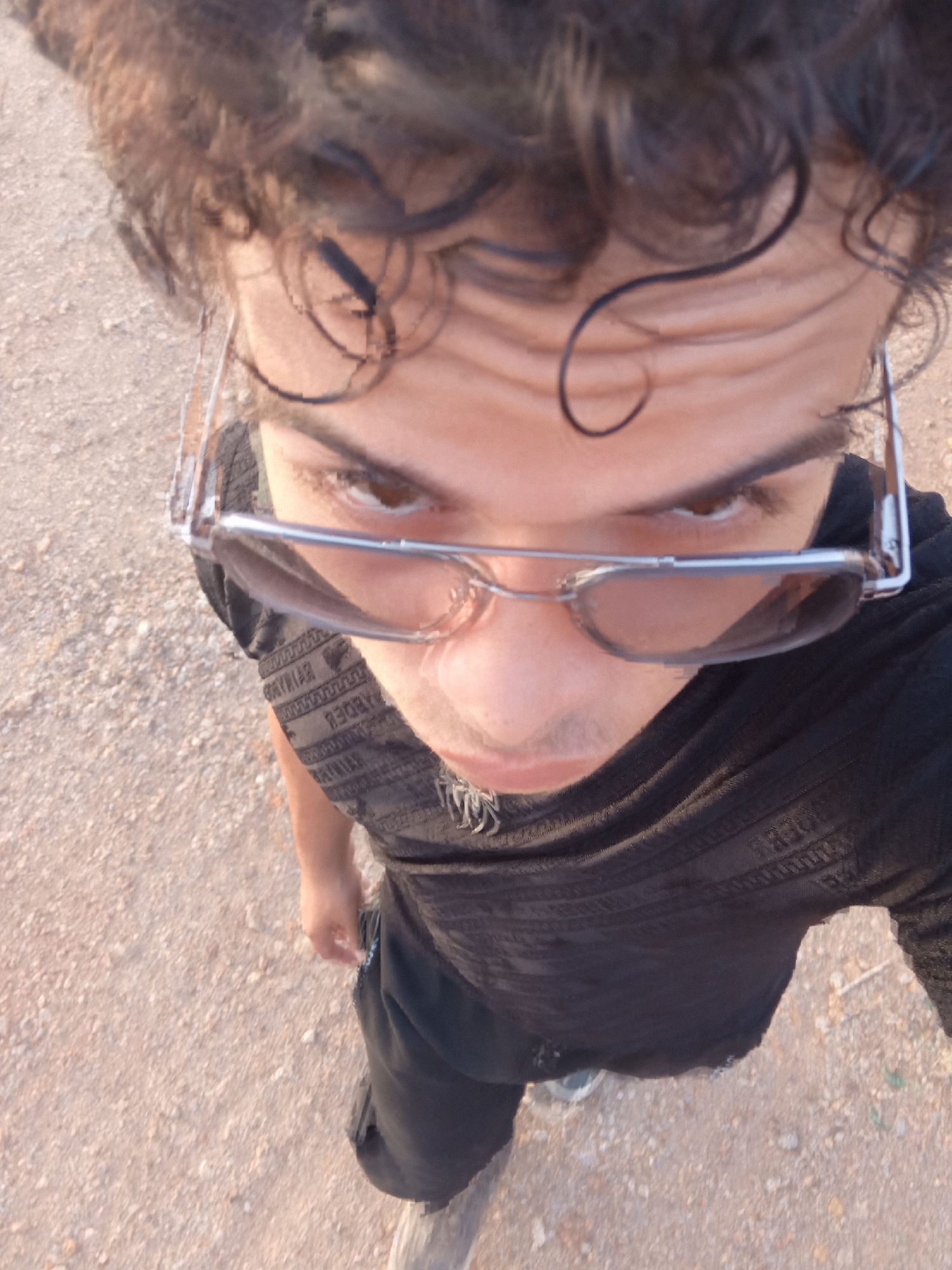 The image captures a high-angle, downward-looking view of a person outdoors in Juan Griego, Venezuela. The subject, likely male, has dark, curly hair and is wearing sunglasses pushed down their nose, revealing their eyes looking up towards the camera. Their expression is neutral, engaged in posing for the photograph.
The person is dressed in a dark, possibly black, short-sleeved t-shirt with a repeating pattern of stylized text or symbols across the fabric, particularly visible on the chest and shoulder. The text appears inverted from the viewer's perspective and is difficult to decipher definitively, but seems decorative, resembling a Greek-like or intricate script. They are also wearing dark pants, and a small chain or necklace is visible beneath the t-shirt's collar.
The setting is an unpaved outdoor area with light brown or tan ground, composed of dirt and small gravel or stones. The ground appears somewhat uneven. The bright, natural lighting suggests it is daytime, with no strong shadows, indicating a bright day. The photograph is taken from directly above the subject, capturing them from head to approximately mid-thigh.
