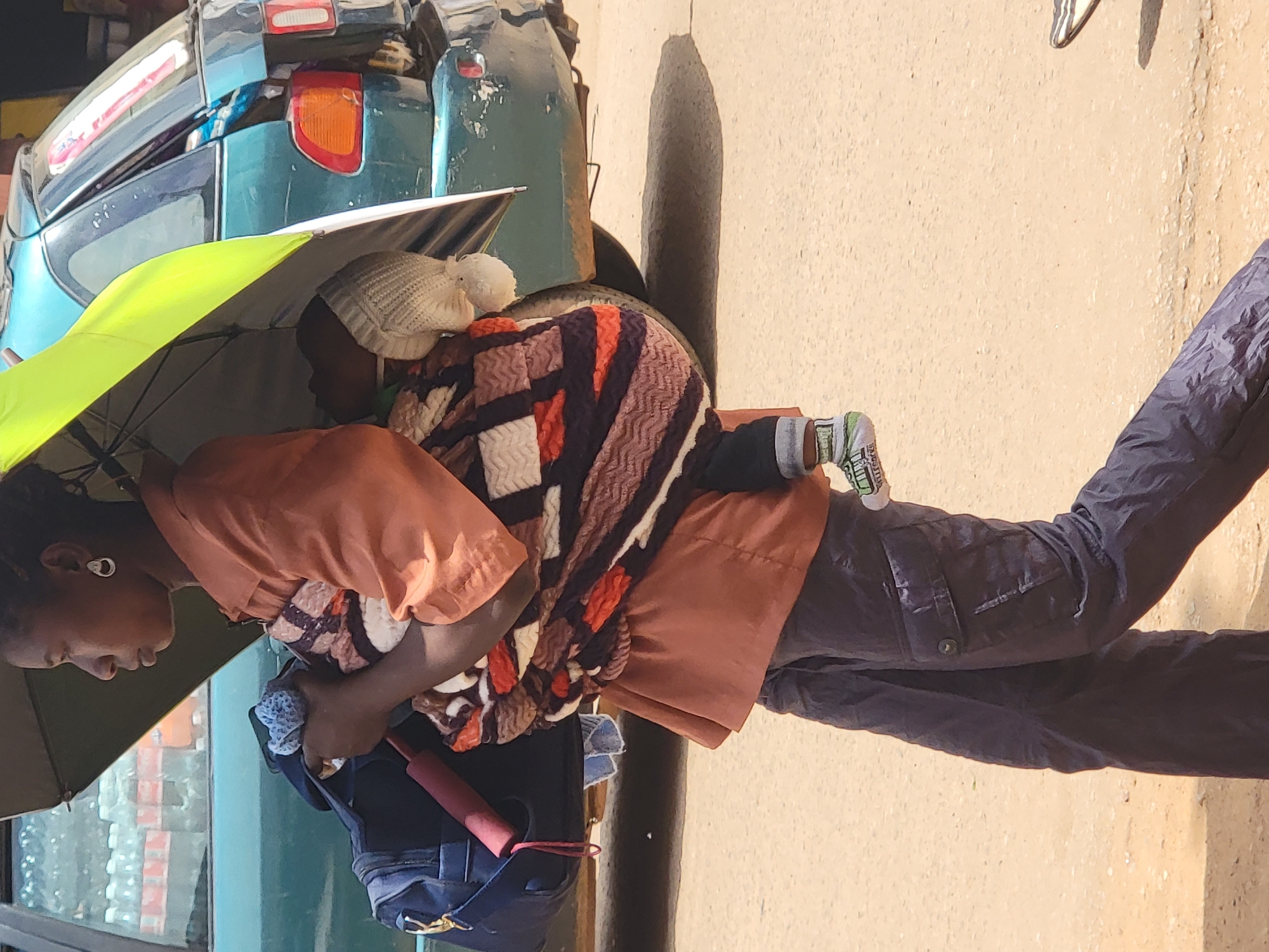 In Maiburiji, Nigeria, a woman stands outdoors, shielded by a green umbrella from the sun. She carries a baby wrapped in a patterned blanket on her back. She wears an orange shirt and dark pants. Behind her is a green car, and a building with window displays is visible. The ground is paved.