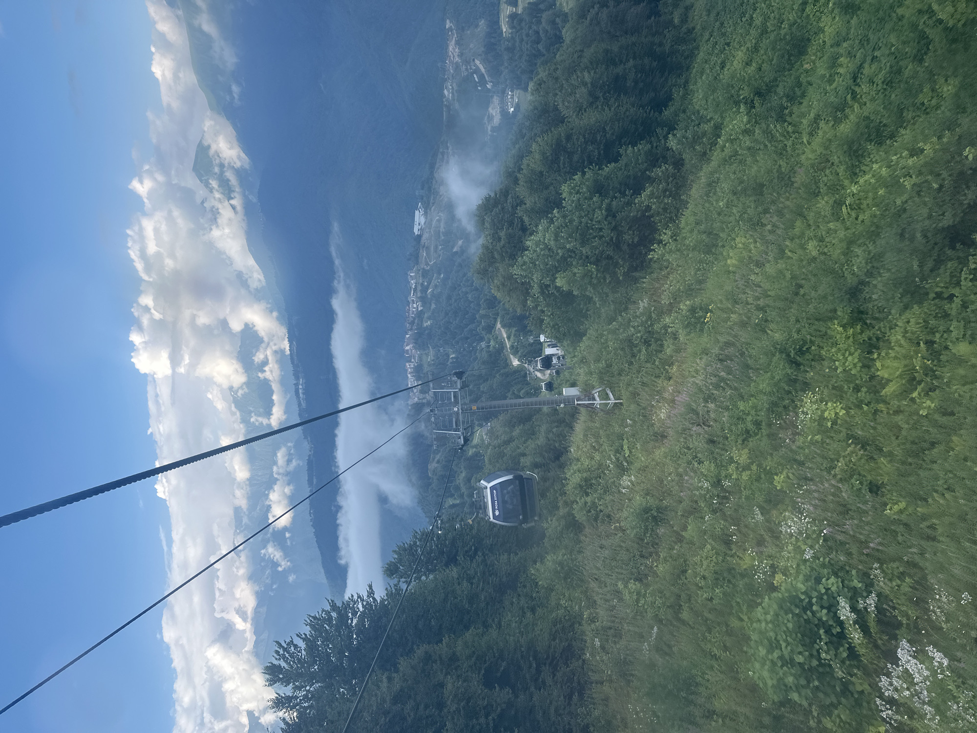 A cable car is ascending a mountain in Estosadok, Russia. The cable car is black and has windows. The cable car is surrounded by lush green trees and vegetation. In the background, there are mountains covered in clouds. The sky is blue and partly cloudy.