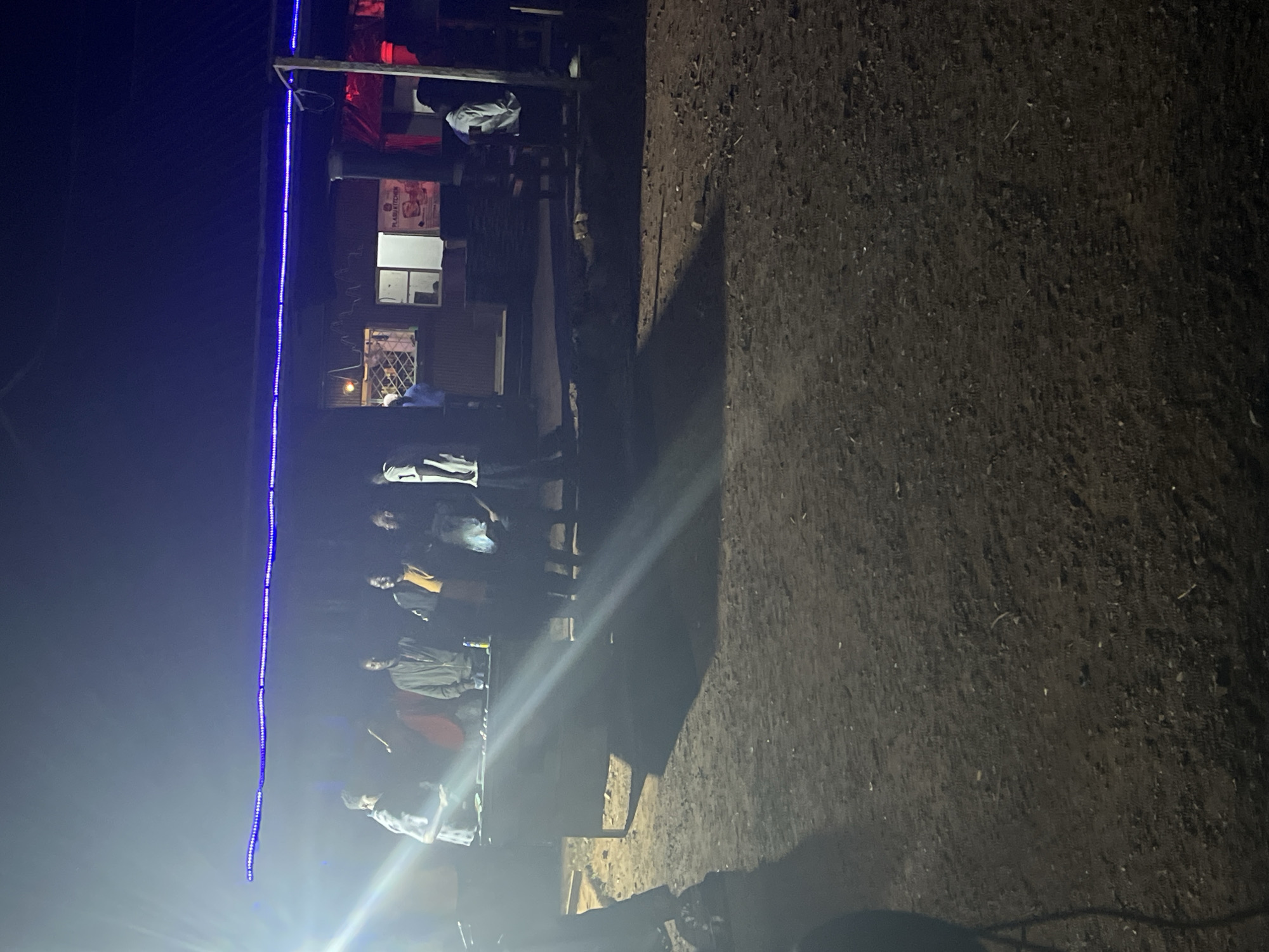A nighttime exterior shot captures a gathering of individuals in a dimly lit, outdoor setting, possibly a market or a local eatery, in Durumi, Nigeria. A blue string of lights illuminates the overhang of a structure behind the group. Several people stand around what appears to be a food stall or counter, with some interacting and others observing. The ground is unpaved, with visible dirt and debris. The overall atmosphere suggests a casual social event or a moment of commerce under artificial lighting.