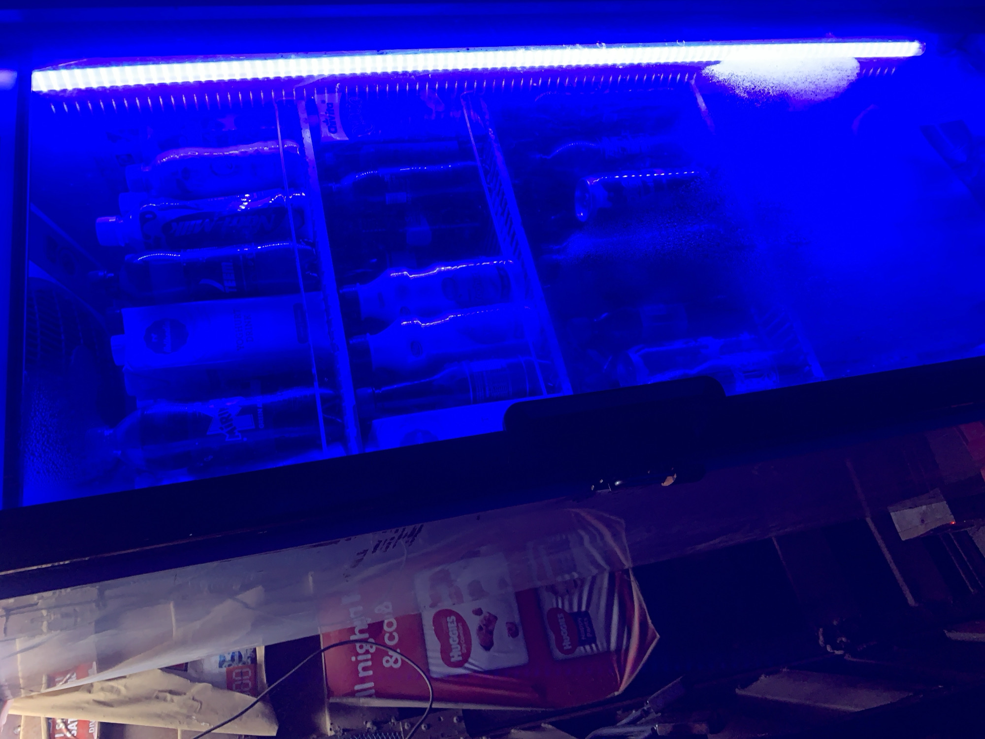 Here is a description of the image:
The image shows a refrigerated beverage display case illuminated with a blue LED light strip. The display case contains various bottles and containers. Beneath the display case are items including a red pack of Huggies diapers and other miscellaneous objects.