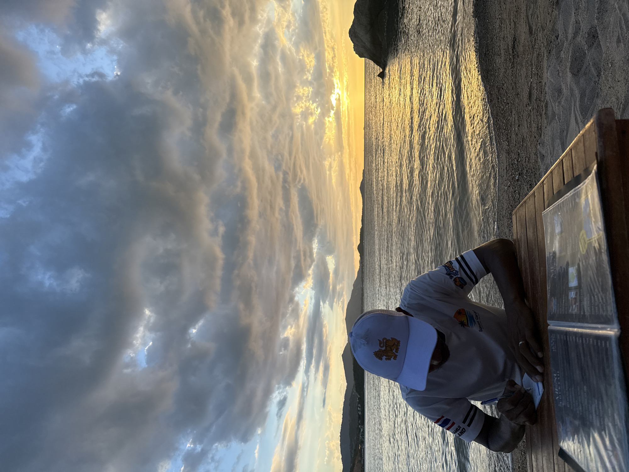 The image depicts a man sitting at a wooden table on a sandy beach in Baan Plai Laem, Thailand. He is wearing a white t-shirt with colorful armbands and a white cap. The man is holding a pen and writing on a piece of paper, with a menu open in front of him on the table. The background features a calm sea with gentle waves reflecting the golden hues of a sunset. The sky is filled with dramatic, cloudy formations, with patches of blue sky and the sun casting a warm glow on the horizon. In the distance, silhouetted hills can be seen. A large rock formation is visible on the right side of the frame, partially submerged in the water. The scene is set during the evening, indicated by the sunset and the soft lighting.