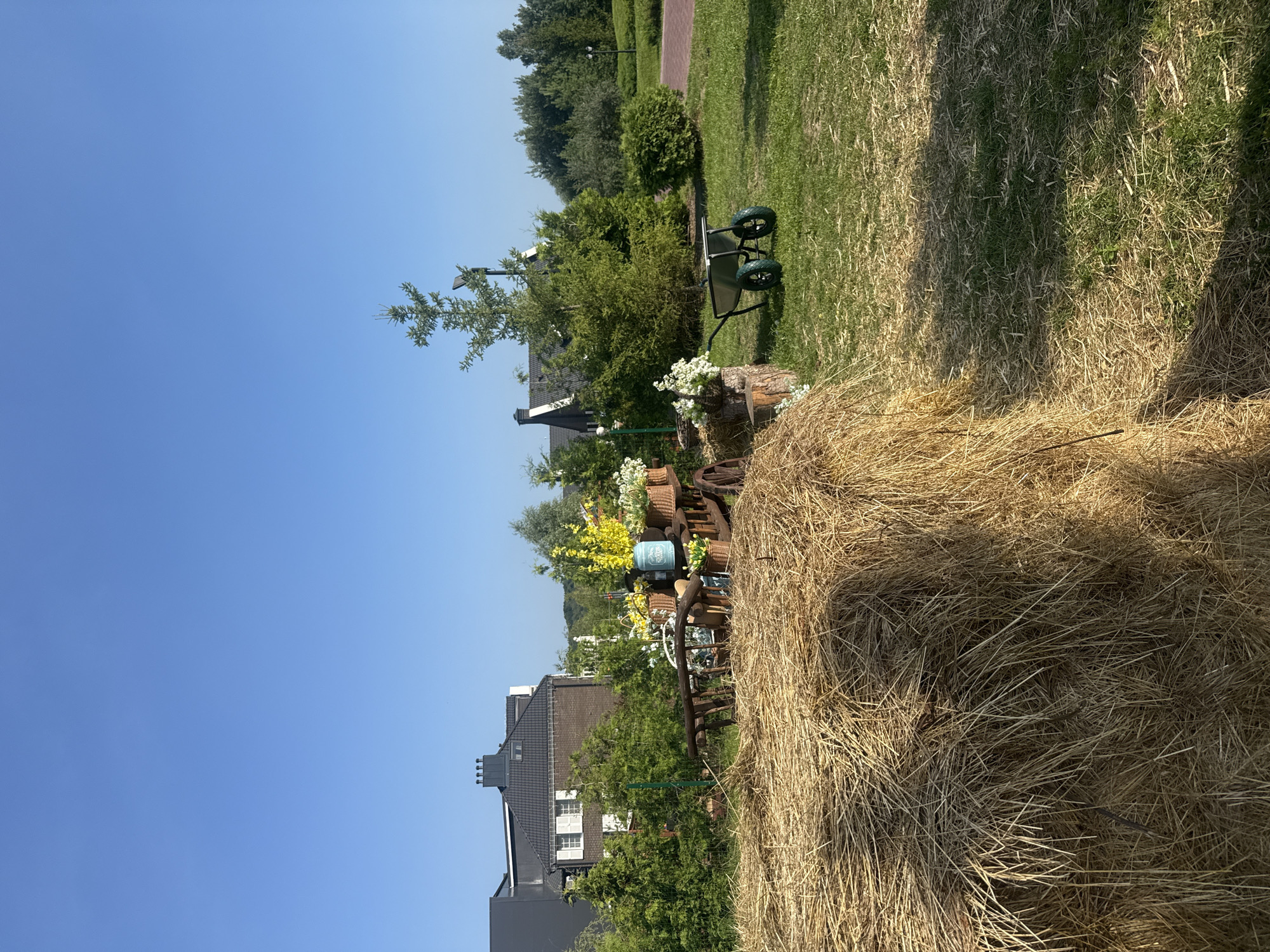 The image depicts a rural-themed outdoor scene on a sunny day. In the foreground, a large bale of dry straw is prominently featured on the left. To the right of the straw bale, an antique wooden cart filled with flower baskets and decorative items sits on a patch of green grass. Among the flowers, there is a blue decorative pitcher. Further to the right, a green wheelbarrow with two wheels is parked on the grass. In the background, several houses with dark roofs and windows are visible, surrounded by lush green trees and bushes. The sky is clear and blue. The location is the city of Seltso, Russia.