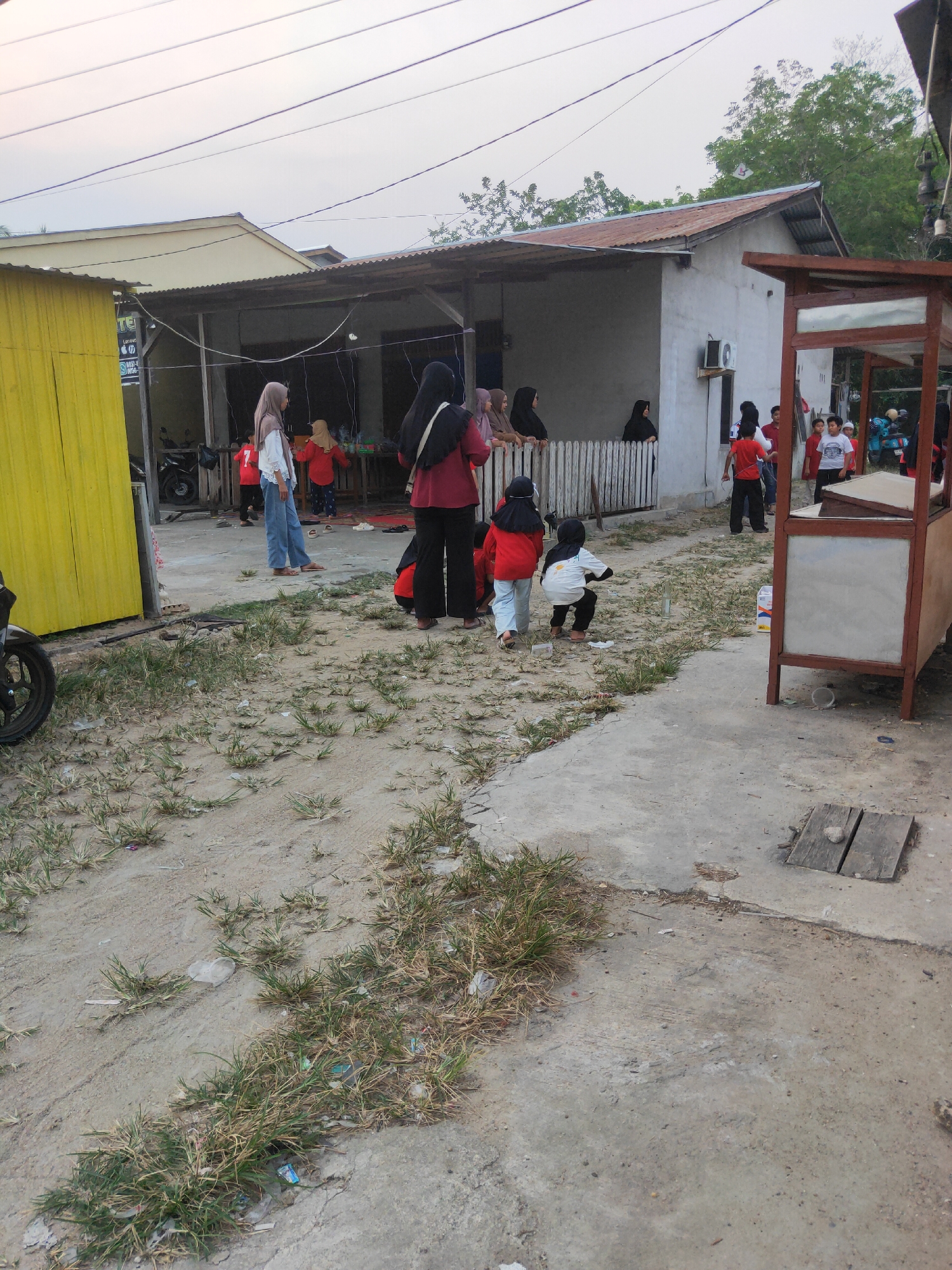 In a scene indicative of a casual gathering in Sintang, Indonesia, individuals are observed in an outdoor setting, possibly a residential area or a communal space. A light-colored building with a corrugated metal roof, typical of structures found in the region, forms the backdrop. To the left, a vibrant yellow corrugated metal structure stands, possibly a shop or an extension of a dwelling.
Several people, predominantly women and children, are present. A woman in a maroon headscarf and top is positioned centrally, appearing to converse with a woman in a light brown headscarf and white shirt. Several children, some in red tops and others in white, are scattered throughout the scene, engaged in various activities. Two children in red are seen near the building, while another child in a white top and black pants is crouched on the ground, seemingly interacting with something unseen. Further to the right, a wooden stall or kiosk with a reddish-brown frame and a white panel suggests a place of commerce or temporary setup.
The ground is a mixture of dirt and concrete, with patches of dry grass and scattered debris, common in the tropical climate of Sintang. Overhead, power lines crisscross the sky, hinting at the presence of infrastructure within the urban or semi-urban environment. The overall atmosphere appears relaxed, with people casually interacting in the outdoor space.