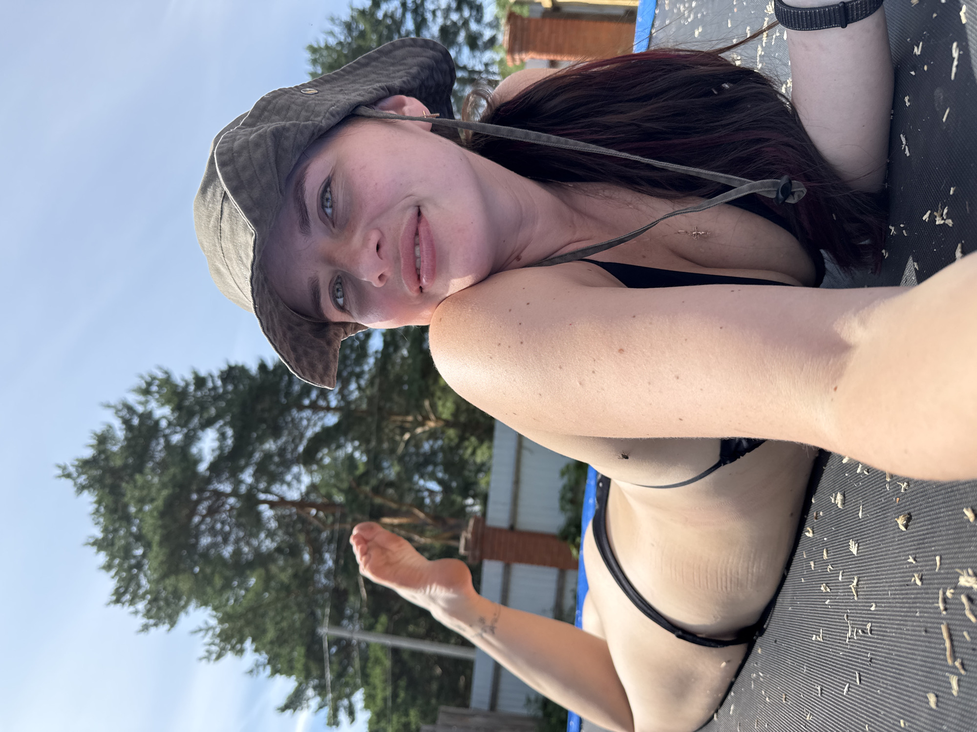A woman wearing a black bikini and a wide-brimmed hat lies on a trampoline outdoors. She is positioned on her stomach, with her head turned towards the camera and a slight smile on her face. Her left leg is extended upwards, revealing a tattoo on her ankle. The background shows trees, a fence, and a clear blue sky. The environment suggests a sunny day, possibly in a rural or suburban setting like Rusalevka, Russia. Small pieces of debris are scattered on the surface of the trampoline.