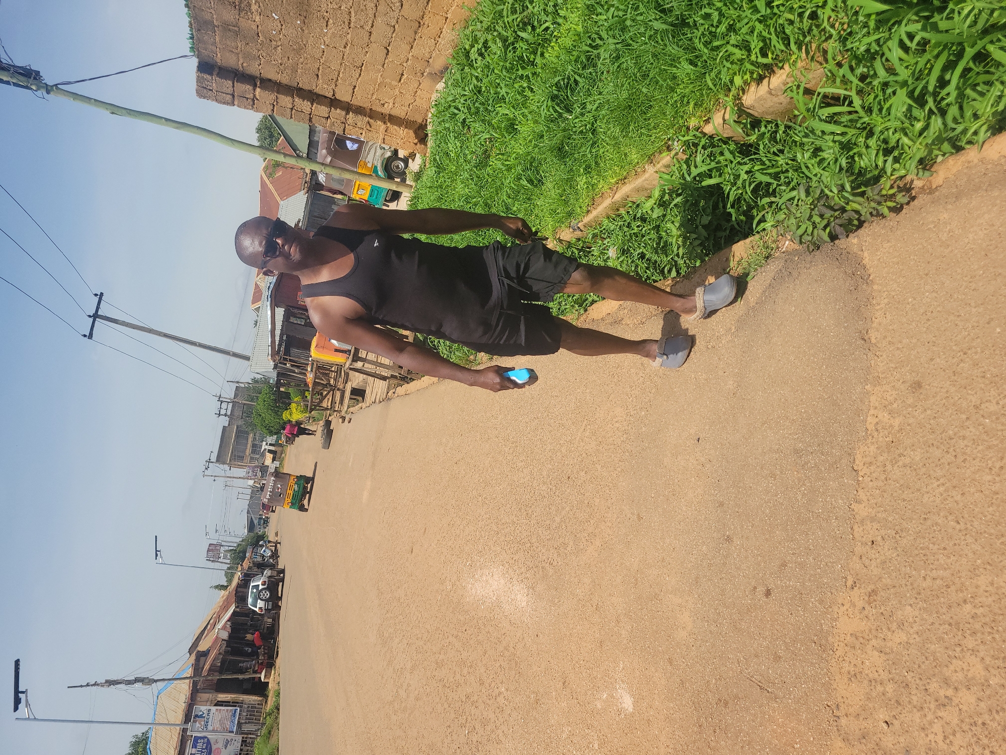 A man wearing a black tank top and black shorts walks on the side of a dusty, paved road in Jos, Nigeria. He is also wearing gray slippers and sunglasses. He holds a light blue object in his right hand. To his left, the road continues, with shops lining the side. A white SUV and a few other vehicles are visible in the distance. To his right, there is a patch of tall green grass and a brick wall. The sky is clear and blue, and there are power lines and utility poles visible throughout the scene. A yellow and green tuk-tuk is driving down the road in the background.