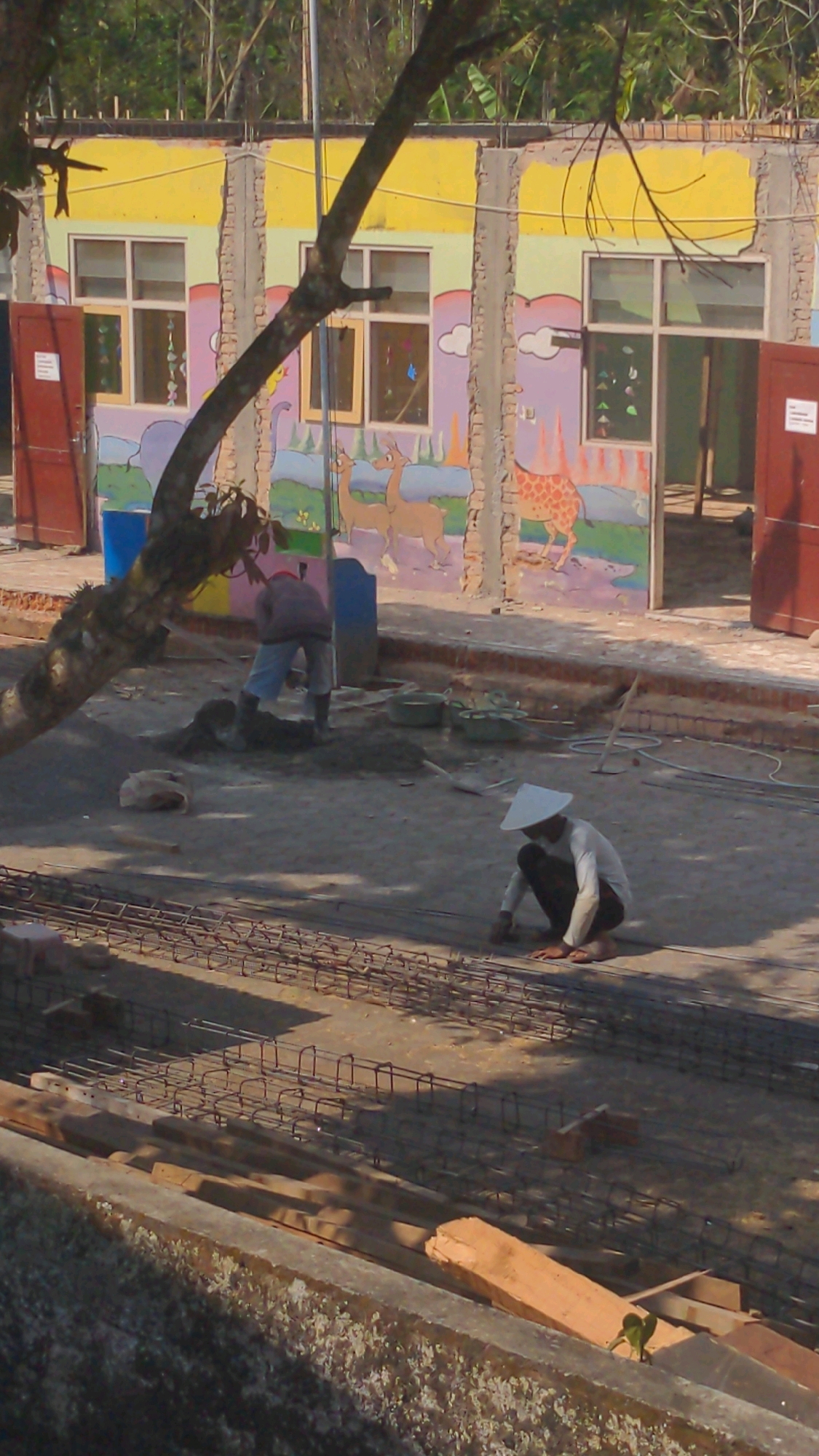 In Tembarak, Indonesia, an outdoor construction scene unfolds. A brightly painted building, likely a school, features murals of animals and clouds on its exterior walls, with yellow and light green paint visible. Several windows punctuate the facade. In the foreground, a man wearing a conical hat and light-colored long-sleeved shirt is crouched down, appearing to work on a rebar grid laid out on the ground. Further back, another construction worker in a reddish-brown shirt and light blue pants is bent over, seemingly mixing or handling cement. The ground is a dusty, compacted surface. Various construction materials, including buckets, tools, and bundles of rebar, are scattered around the site. The overall impression is of a construction project in progress, possibly part of the renovation or expansion of a community building.