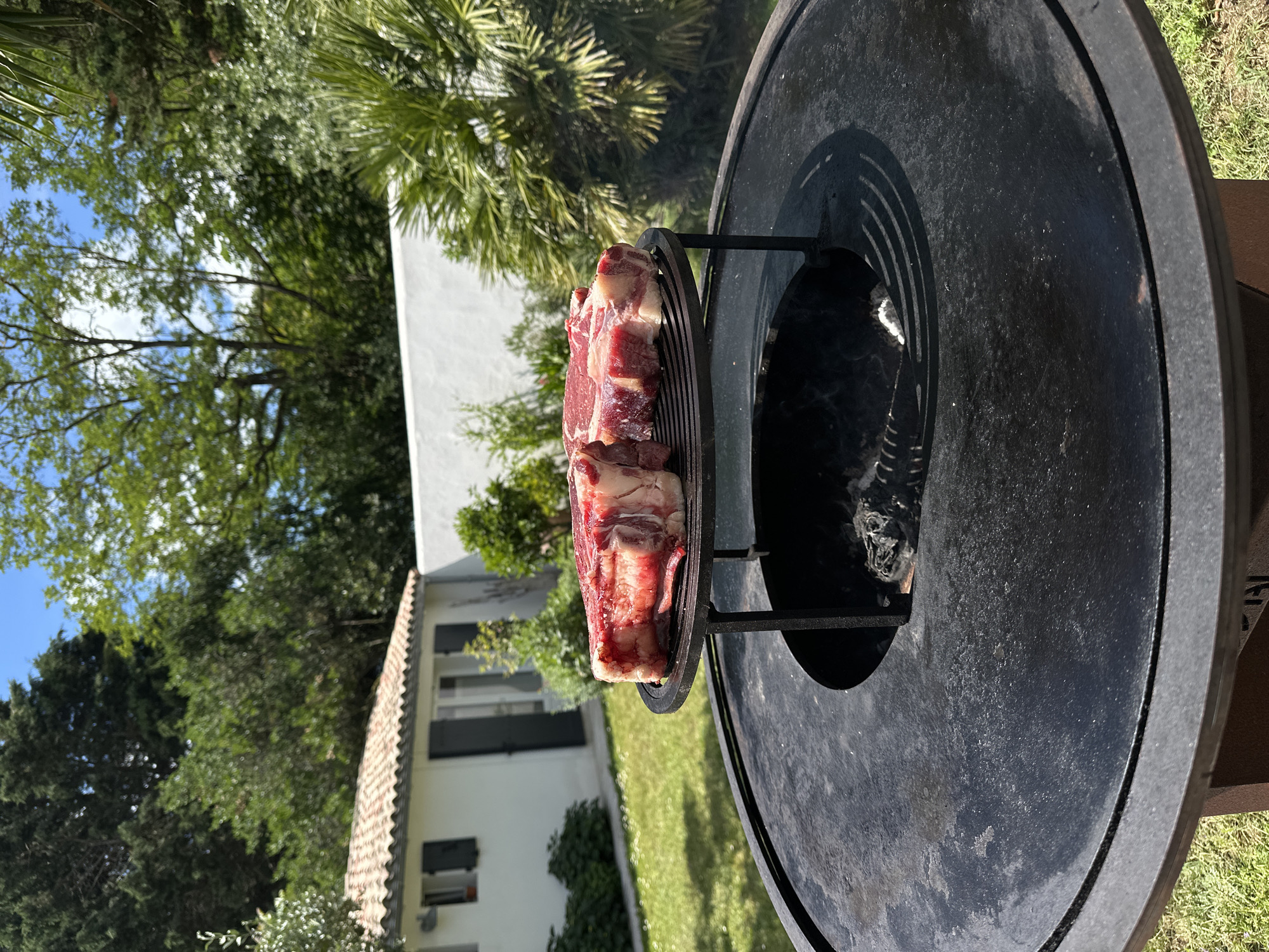 A cut of beef rests on a metal grate inside a grill. The grill is positioned outdoors on a grassy lawn, with trees, bushes, and a two-story house visible in the background. The house is white with a tiled roof. The scene is located in La Couarde-sur-Mer, France.