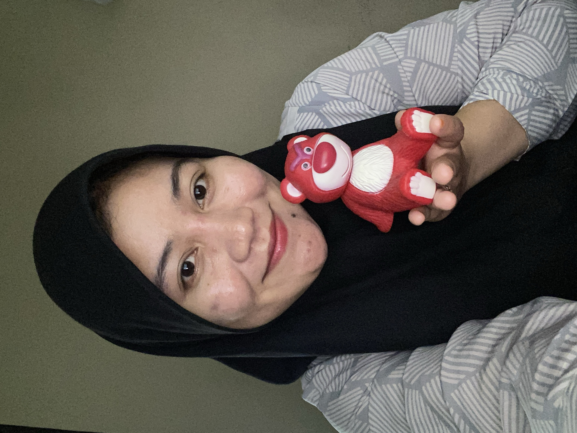 A woman wearing a black hijab and a grey and white patterned shirt holds a red Lotso Huggin' Bear toy in her left hand. She is looking at the camera and smiling. The background is a plain, textured wall. The scene appears to be indoors, possibly in a home. The image is a selfie. The location is not discernible from the image itself.