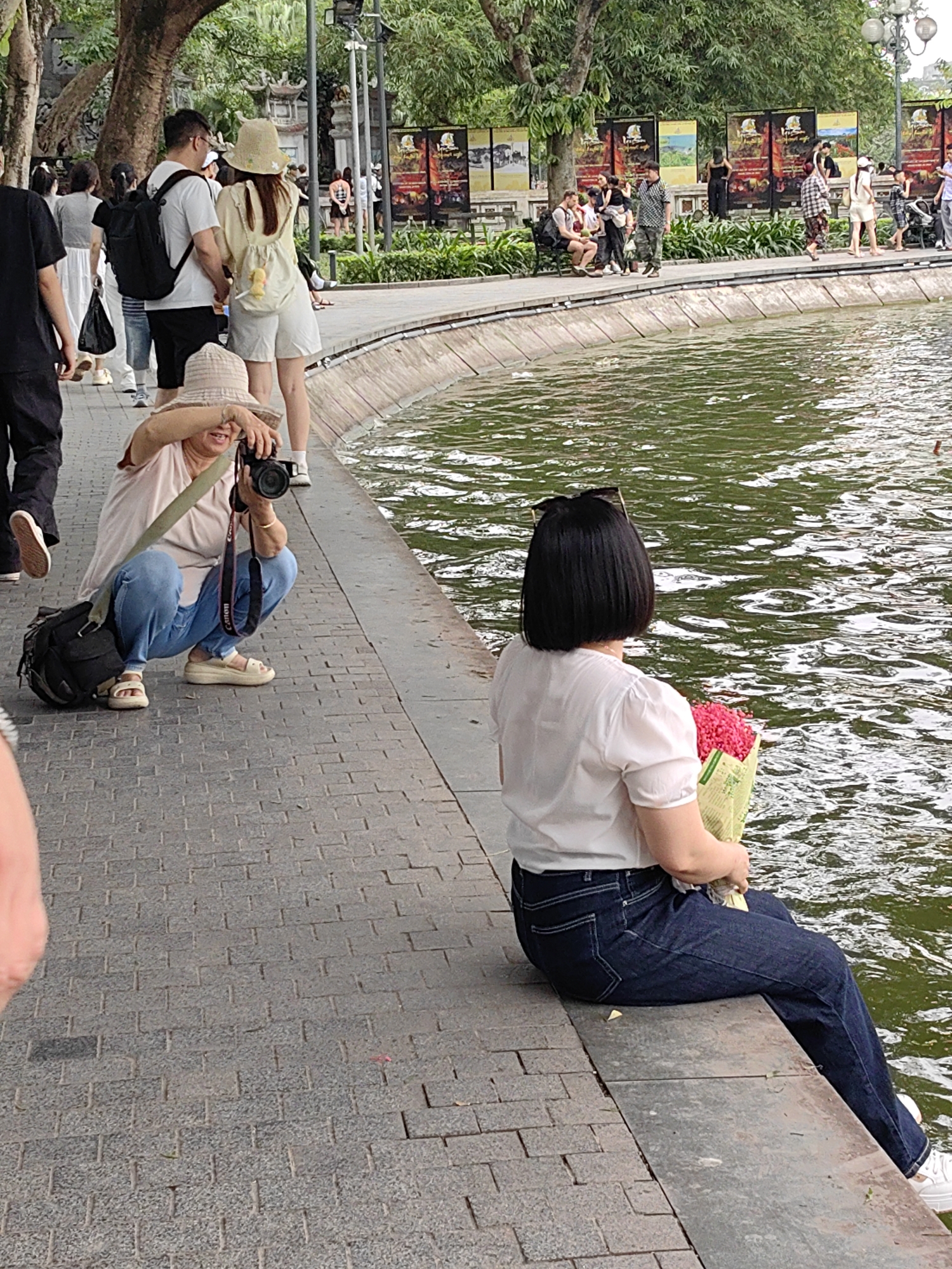 In Hà Nội, Vietnam, an eye-level outdoor medium shot captures a scene next to a pond with a concrete retaining wall. A woman with short black hair and sunglasses perched on her head sits on the wall, her back to the camera. She wears a white short-sleeved shirt and dark blue jeans, holding a bouquet of red flowers wrapped in pale green paper.
To her left, a woman wearing a khaki sun hat, light pink t-shirt, and blue jeans is crouched down, aiming a black Canon digital camera towards the pond. A black camera bag rests next to her. Other pedestrians stroll behind the woman with the camera, including a couple, one with a backpack.
The pond's surface is slightly rippled, reflecting the sky. Benches and streetlamps line the walkway behind the pond. Posters are displayed on a wall in the background. The day appears to be overcast.