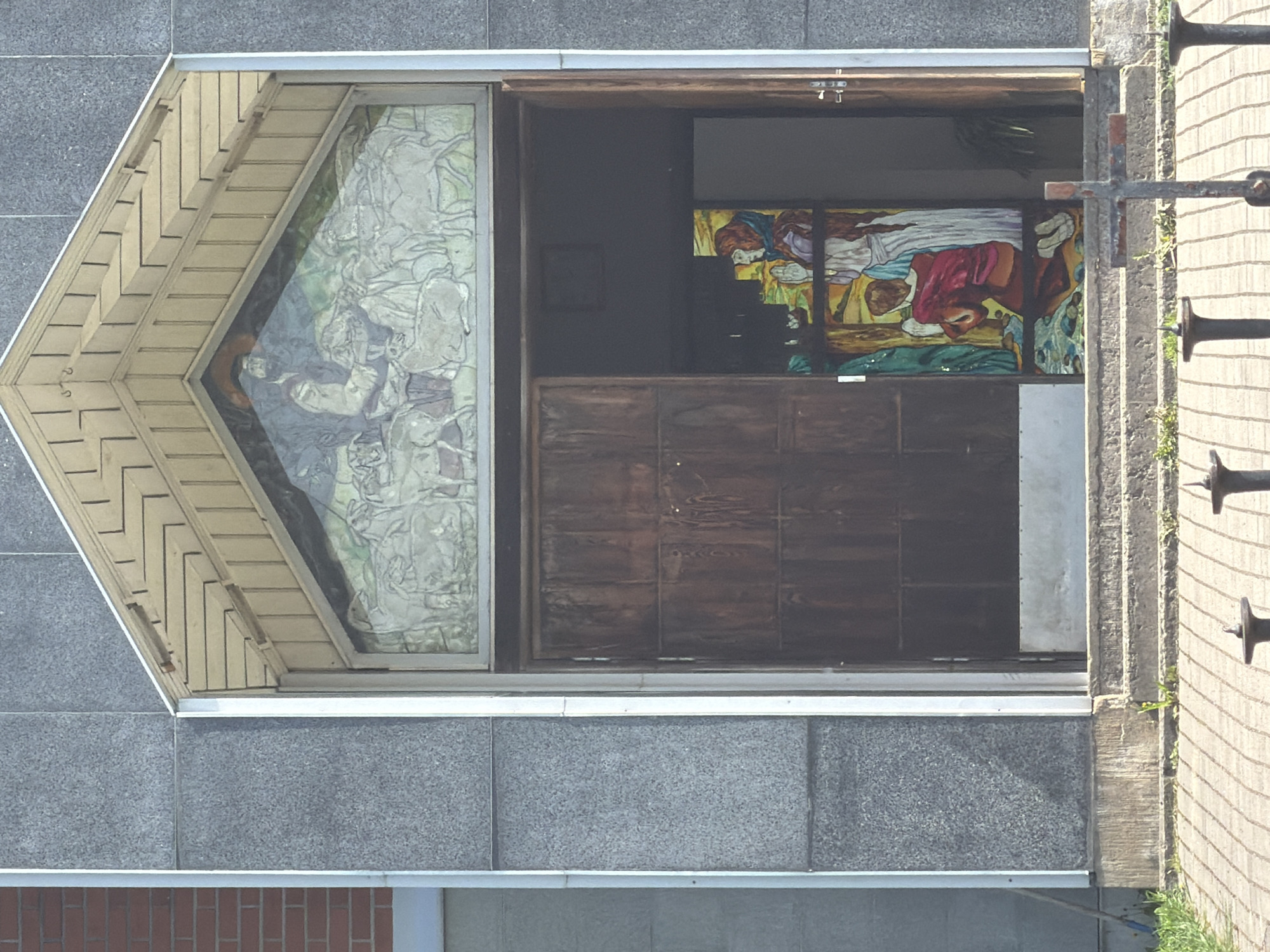 An eye-level, full shot shows a partial view of a building, including a window with a beige, triangular awning above a square section filled with a stained-glass mural; next to it is a dark wooden door panel, a narrow doorway and a wall with metal crosses. The building is made of gray, granite-like stone blocks; a lower section has tan-colored bricks. There is no indication of the location in Tokkari, Russia.