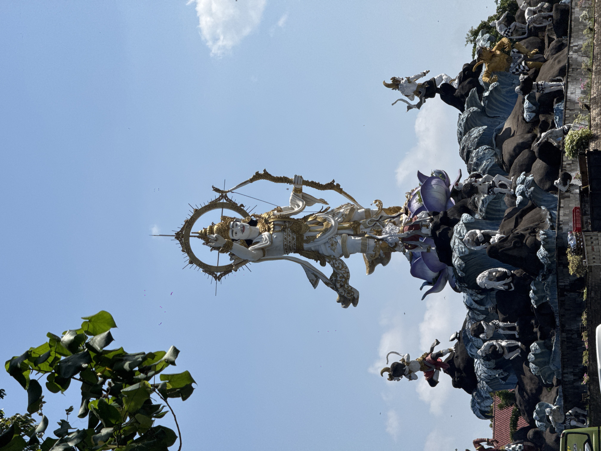 The image depicts the colossal statue of Lord Vishnu on Garuda, a prominent landmark in Denpasar, Indonesia, specifically located at the junction of Jl. Letda Tantra and Jl. Uluwatu. The central figure is a majestic statue of Lord Vishnu, adorned in white and gold, holding a bow and arrow, signifying his divine prowess. He stands atop a vibrant purple lotus, which itself rests on a complex, multi-tiered structure resembling a mountain or a rocky outcrop.
This base is intricately decorated with numerous smaller statues of mythical creatures and figures. On the left, a statue of a monkey-like deity is positioned, with another resembling a warrior further to the right. The lower levels of the structure are populated with various white statues, some appearing to be human-like figures in various poses, and others with animalistic features. The entire monument is surrounded by a decorative representation of waves, creating a dynamic and dramatic visual effect.
The statue is illuminated by bright sunlight, casting clear shadows. The background consists of a clear blue sky with a few scattered clouds, and a branch of a leafy green tree hangs into the frame from the top left. The overall scene is one of grandeur and religious significance, a testament to Balinese artistry and spirituality.