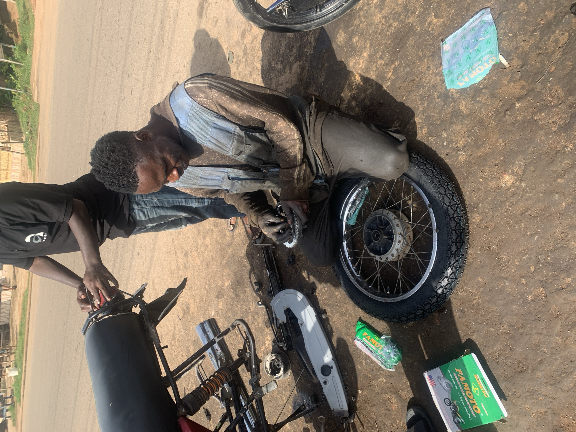 In Gura Top, Nigeria, a mechanic is seen working on a motorcycle. The man, dressed in oil-stained overalls, is kneeling on the ground, his hands engaged with the motorcycle's chain and sprockets. The motorcycle is partially disassembled, with parts scattered around him. A second person, wearing a black t-shirt, stands nearby, possibly assisting or observing.
The scene appears to be outdoors, on a paved surface that looks like a road or a workshop area. The bright sunlight casts strong shadows, indicating it's likely midday. The ground is dusty and dirty, characteristic of a mechanic's workspace. In the background, a line of trees and some simple structures can be seen, suggesting a rural or semi-urban setting.
Scattered on the ground are various items related to motorcycle repair, including a new motorcycle chain in a green package labeled "FAMOTO" and what appears to be a box of spare parts, also with "FAMOTO" branding. A crumpled piece of paper, possibly packaging or instructions, is also visible. The overall impression is one of diligent work in a practical, perhaps humble, environment. The mechanic appears focused on the task at hand.