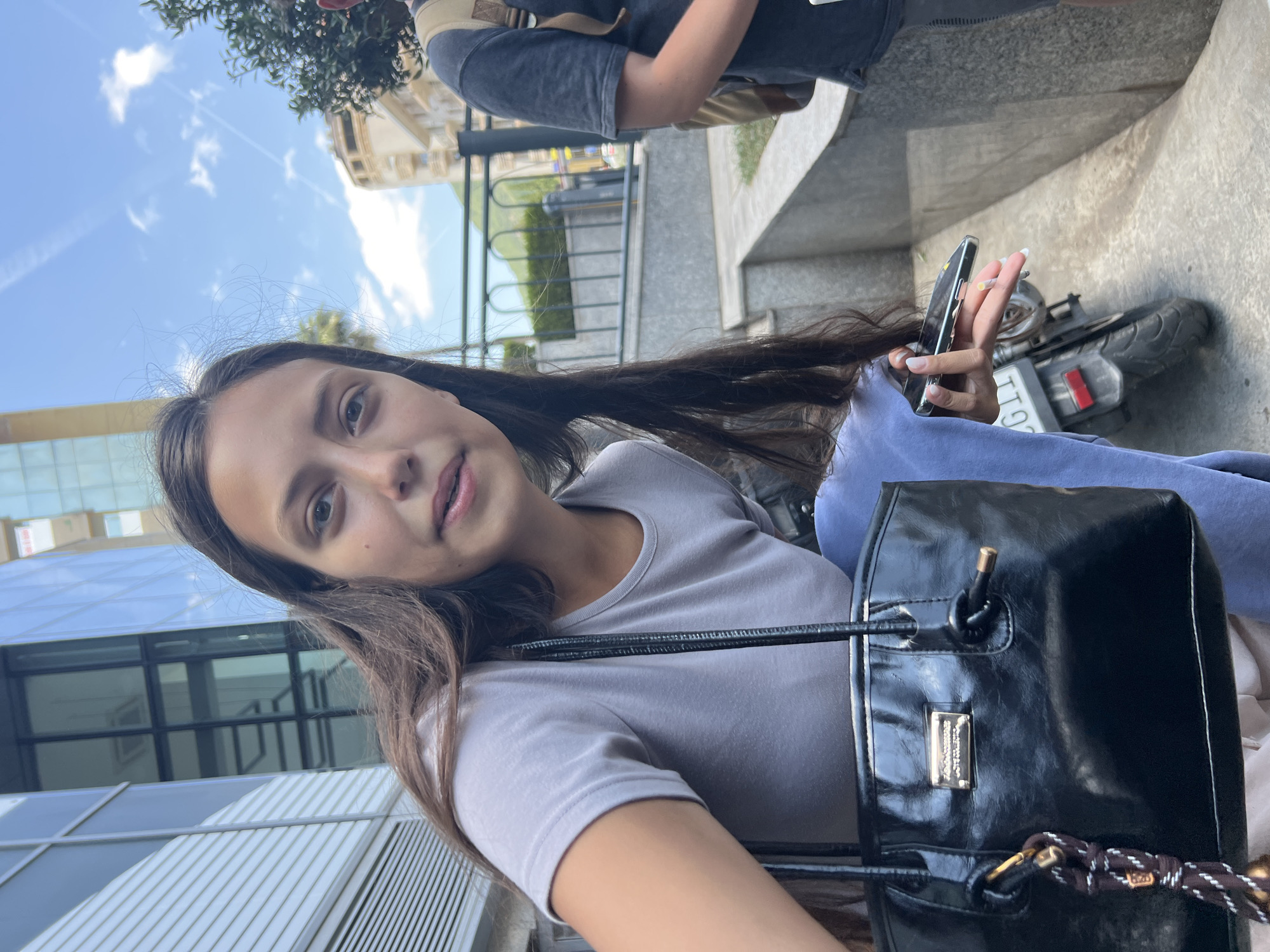 A young woman with long, dark hair and a light gray t-shirt is captured in a close-up, slightly angled shot. She holds a smartphone in her right hand, and a black, shiny tote bag hangs from her left arm, with a decorative tassel attached. Behind her, a parked motorcycle is partially visible, along with a portion of another person wearing a backpack and dark t-shirt. The background suggests an urban environment in Budva, Montenegro, with modern buildings, a decorative fence, and lush greenery. The sky is blue with some white clouds and contrails. The lighting indicates it is daytime, likely late morning or early afternoon.