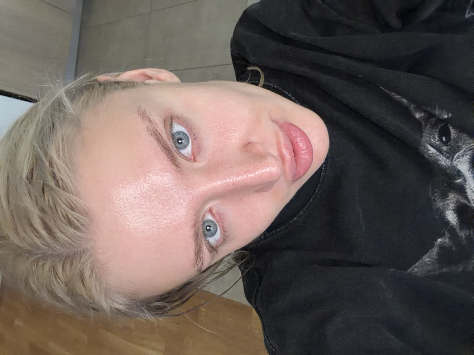 The image displays a high-angle, close-up portrait of a fair-skinned individual, likely female. The subject has blonde hair, appearing wet or styled, with a visible central parting. Features include light blue eyes, natural-toned eyebrows, and full, pink-tinted lips. The skin exhibits a subtle sheen. The individual is wearing a dark, possibly black, crew-neck top with a partial graphic design on the lower chest, depicting the head of an animal with visible antlers or horns. The background reveals an indoor setting with two distinct flooring types: light brown wooden planks are visible to the upper left, and light grey tiles are present to the upper right, separated by a dark vertical line, possibly an architectural joint or door frame. The specific location within the city of Belgrade, Serbia, cannot be determined from the visual information provided in the image.