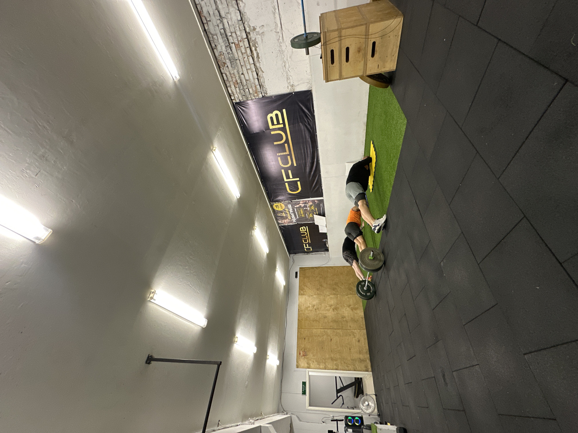 The image shows an indoor CrossFit gym. The location is likely Chaikovsky, Russia, as indicated by the "CFCLUB" banner and other Russian-language signage visible in the background. The gym features black rubber floor tiles and various pieces of fitness equipment, including a wooden plyometric box, barbells with weight plates, and artificial turf. Two people are engaged in weightlifting. One is bending over a barbell, presumably preparing to lift it. The other is nearby, likely spotting. The gym has fluorescent lighting fixtures and a wall with a "CFCLUB" banner displayed.