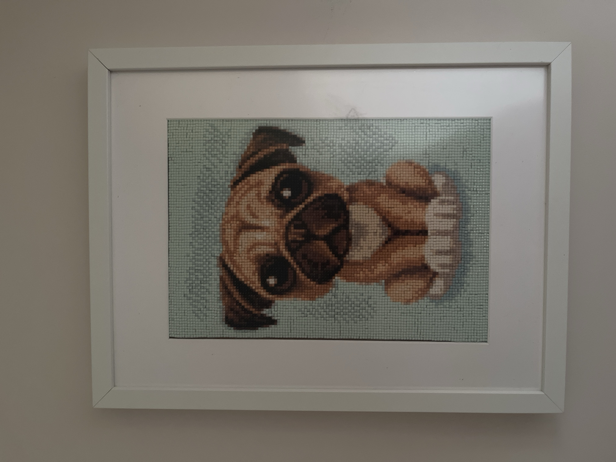 A framed cross-stitch image of a pug dog is hung on a light-colored wall. The pug is depicted sitting and facing forward, with large, dark eyes and a light brown coat. The background of the cross-stitch is a pale blue grid pattern. The frame is white and simple in design. The image does not contain any information about the city of Haabneeme alevik, Estonia.