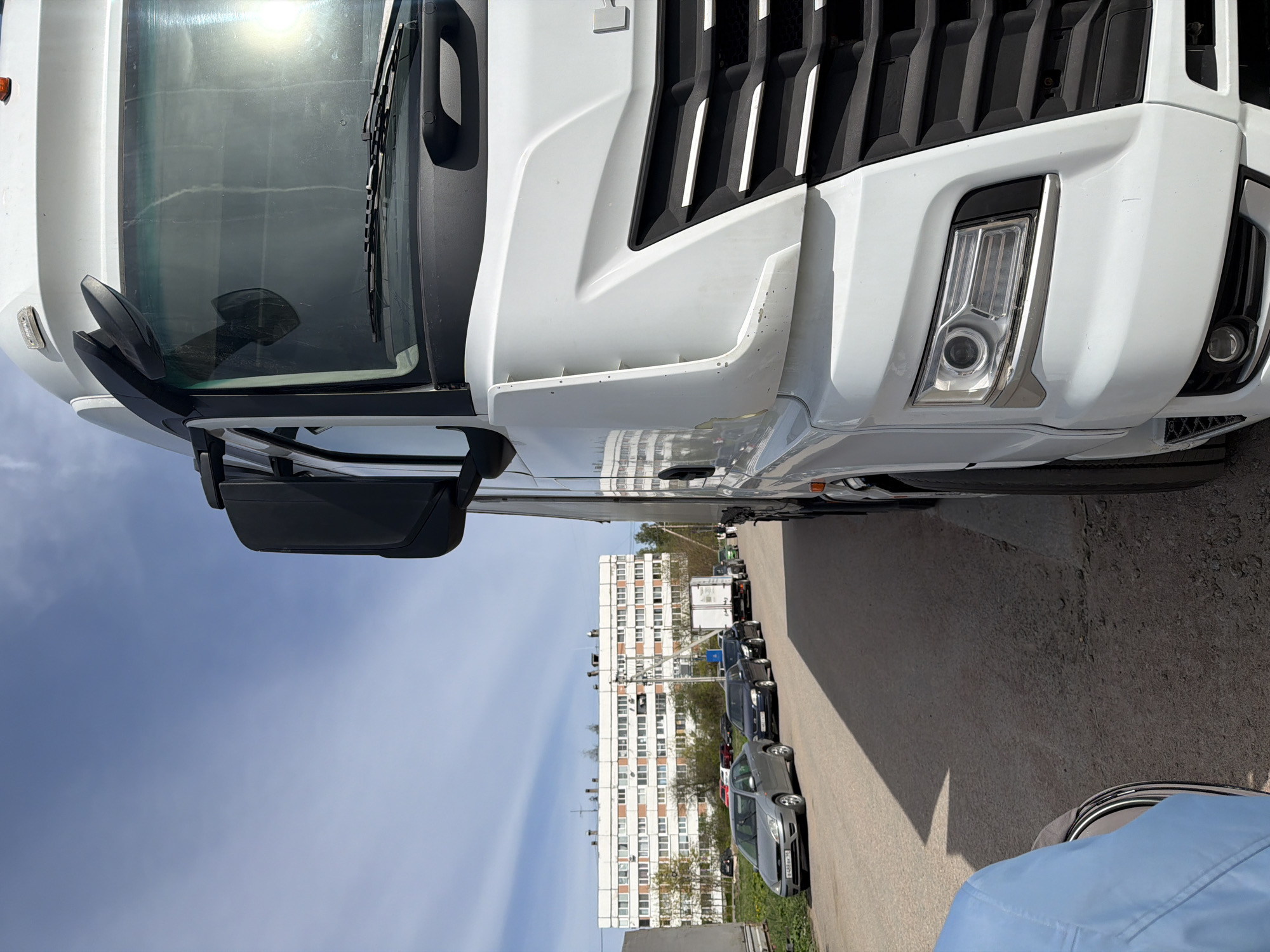 The image shows the front of a white truck parked on a street lined with parked cars. In the background, there is a tall apartment building with multiple windows. The sky is blue with some clouds. The image appears to be taken in Tavry, Russia, judging from the license plates of the parked cars.