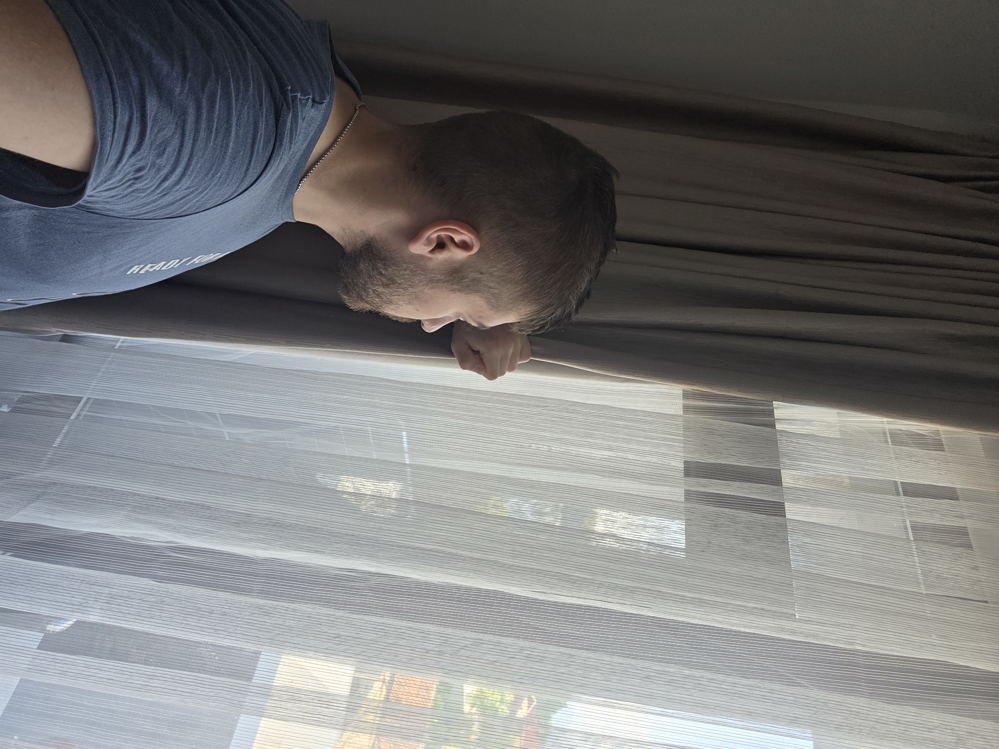 A man is positioned near a window. He has short, dark hair and a beard, and is wearing a blue t-shirt. A silver chain is visible around his neck. His face is close to the window, and his hand rests on the edge of the window frame. The window is covered with white blinds, and a set of dark curtains is above the blinds. Outside the window, a partial view of buildings and greenery suggests an outdoor setting. The location in Yagodovo, Bulgaria is not able to be determined based on the image.