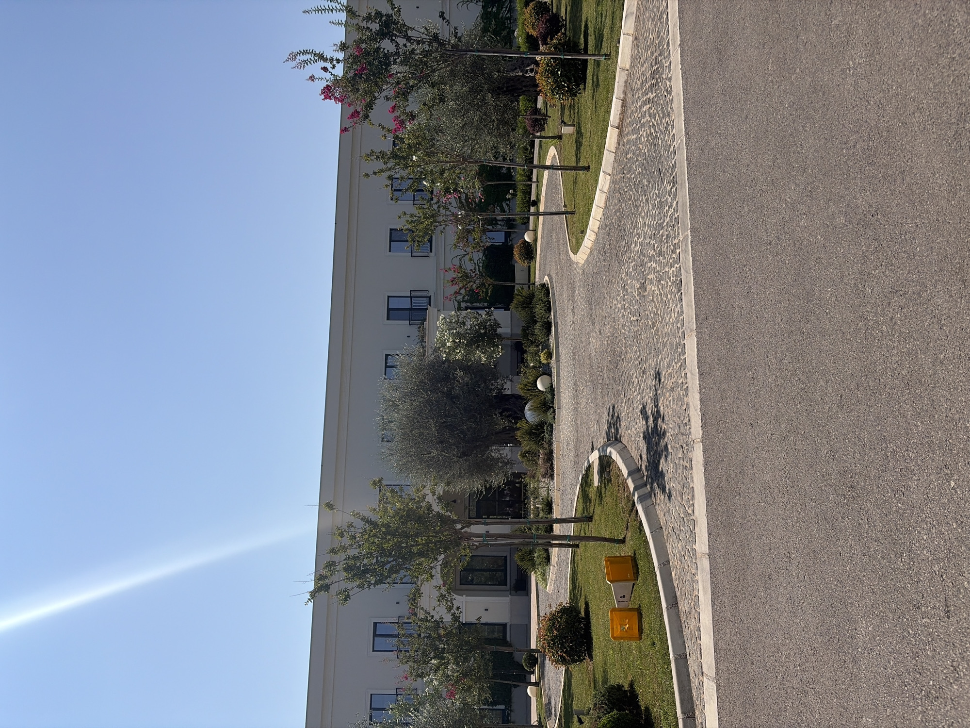 A long, modern, cream-colored building with a flat roof stands under a bright, cloudless blue sky. Sunlight streams down from the upper left, creating a bright glare that obscures a portion of the sky. The building has multiple windows with dark frames and small balconies. In front of the building, a circular driveway paved with cobblestones curves around a landscaped area. This area contains neatly trimmed green grass, various bushes, and several small trees. Some of these trees have pink flowers. Two large, white spherical decorative objects are visible in the landscaping. On the grass, near the edge of the cobblestone driveway, are two yellow traffic cones. The foreground is dominated by an asphalt road. The scene appears to be an entrance to a hotel or resort in Podgorica, Montenegro.
