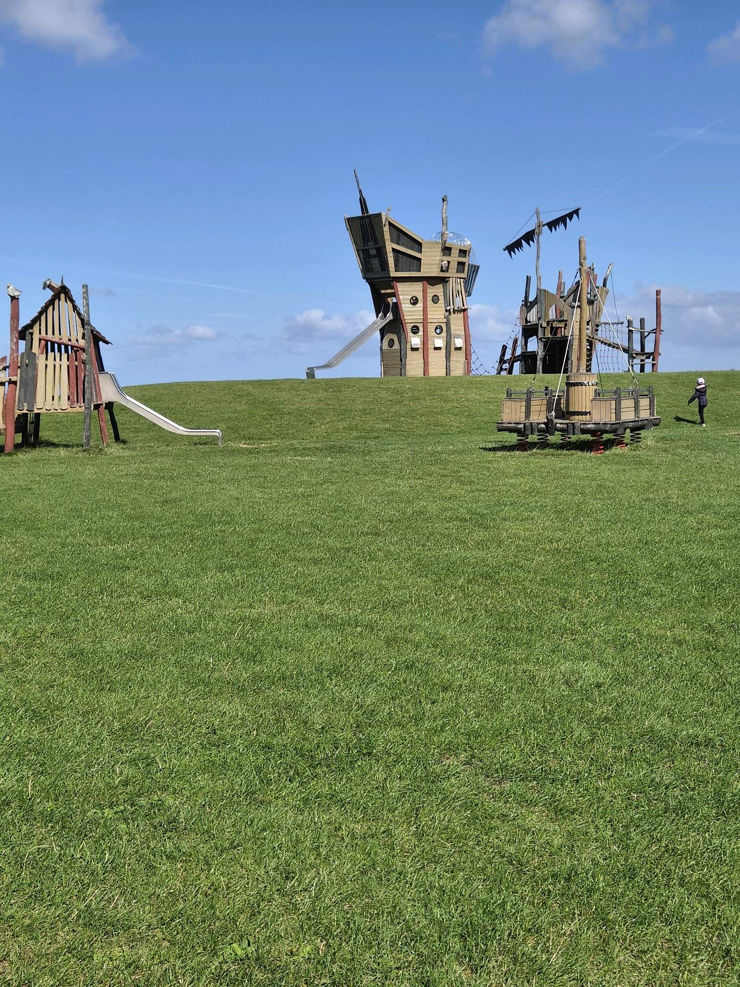 The image depicts a sun-drenched outdoor playground on a grassy hill under a bright blue sky with scattered white clouds. In the middle ground, a large, wooden, ship-themed play structure dominates the scene, featuring multiple levels, slides, and a net climber. To the left, a smaller, rustic wooden fort with a slide is visible. In the right middle ground, a small, red and wooden spring rider toy is positioned. In the far right of the image, a solitary child, wearing a dark jacket with a pink hood and dark pants, walks away from the playground structures. The environment is clearly a public park or recreational area, likely in Dorum, Germany, given the context provided. The grass is lush and green, indicating a well-maintained area.