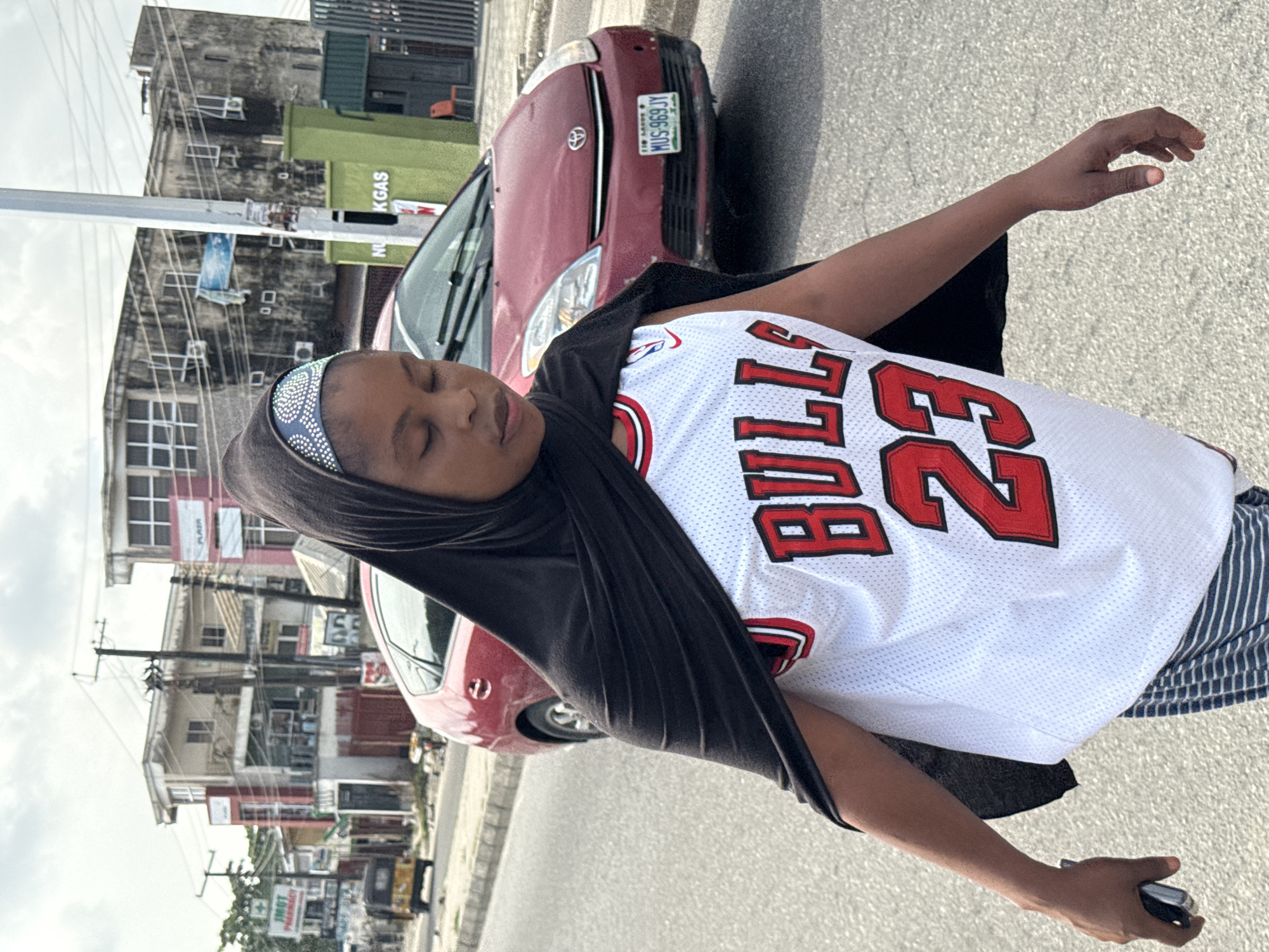 An upright, daytime shot captures a person on a street in what appears to be an urban setting in Lagos, Nigeria. The central figure, likely female, has her eyes closed and is wearing a white Chicago Bulls basketball jersey with "BULLS" and the number "23" printed in red. She also wears a black long-sleeved shirt underneath, striped pants, and a dark head covering with a light blue, sequined band across the forehead. A black mobile phone is held in her right hand.
In the background, a maroon Toyota sedan is visible, bearing a Nigerian license plate "MUS-969 JY LAGOS". The street features utility poles with numerous overhead wires, and a mix of multi-story buildings, some appearing weathered. Among the visible signs on the buildings are "PLAZA" and "IMOT PHARMACY." A prominent green rectangular structure, possibly a commercial container, displays text that includes "NUN KGAS". The bright, even lighting suggests it is daytime. The scene conveys a casual, everyday moment in a busy city.