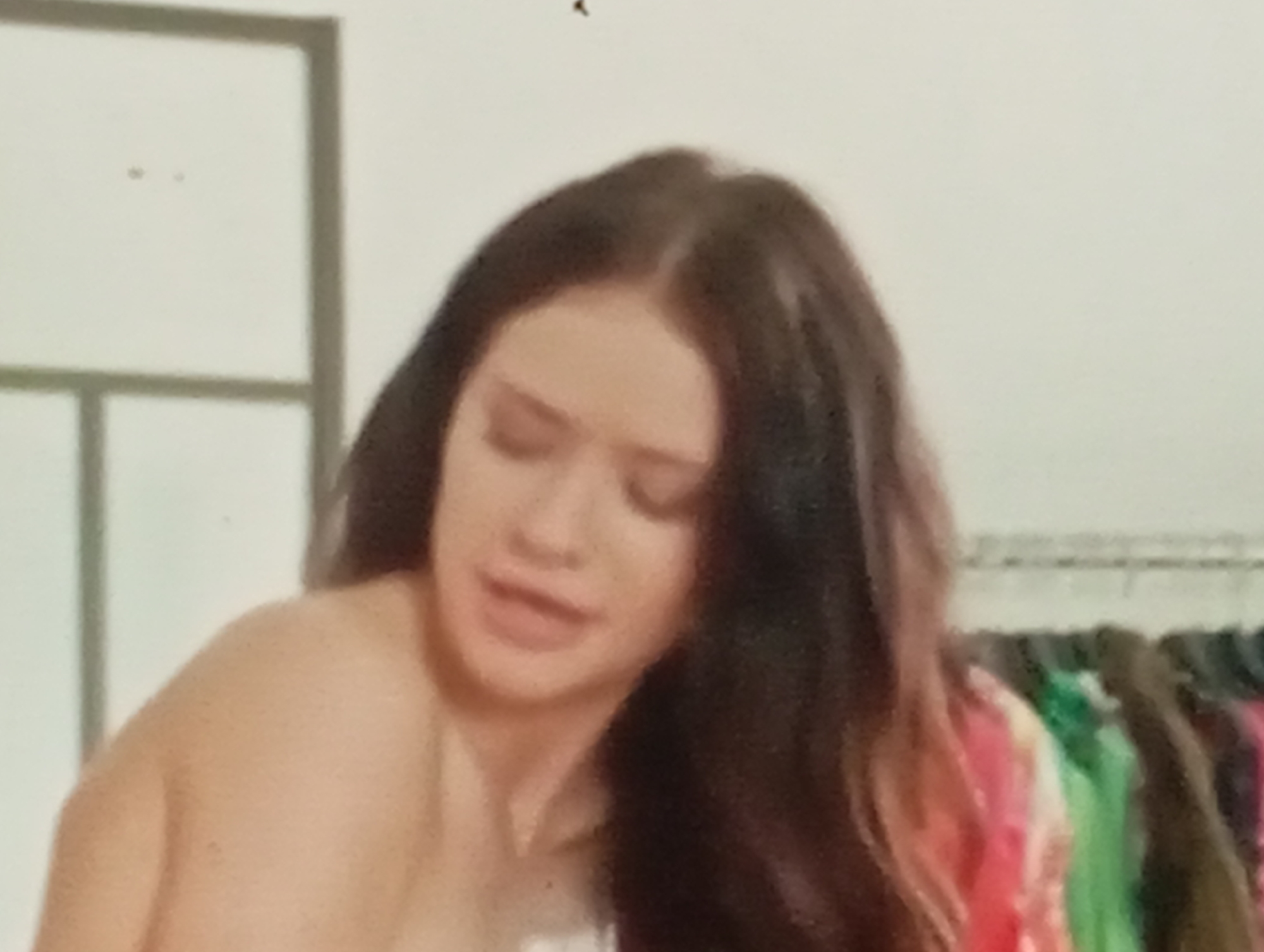 The media file shows a close-up, low-resolution, portrait of a young woman with dark, long hair. She is facing slightly away from the camera, with her head tilted downwards and her eyes closed. Her expression appears to be one of discomfort or distress, with her mouth slightly open. She is bare-shouldered, and her skin tone is fair. The background is out of focus but appears to be an indoor setting, possibly a room or a changing area. To the right of the woman, there is a rack of colorful clothing, suggesting a retail environment or a personal wardrobe. A section of a light-colored wall with a dark frame is visible on the left. The overall lighting is soft and diffused, with no strong shadows. There is no visible text in the image. The context suggests this image might be from Sofiivska Borshchahivka, Ukraine.