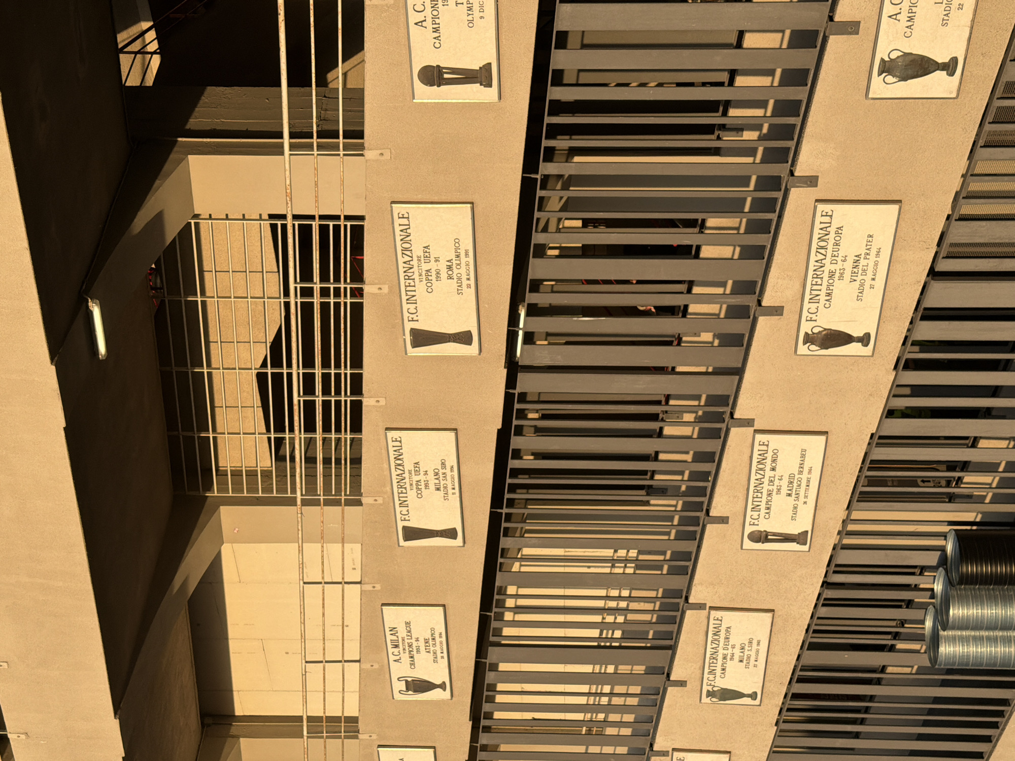 This image depicts a section of wall adorned with plaques commemorating the achievements of the Italian football clubs A.C. Milan and F.C. Internazionale. The plaques are displayed on a building exterior, partially obscured by a metal grate. The plaques detail specific titles won, such as Champions League and UEFA Cup victories, along with the respective year, city, and stadium where the match took place. Given the clubs listed, the location is likely somewhere in Milan, Italy, the home city of both teams. The building has a combination of concrete and metal elements and some kind of ventilation. The scene appears to be in a low-lit area.