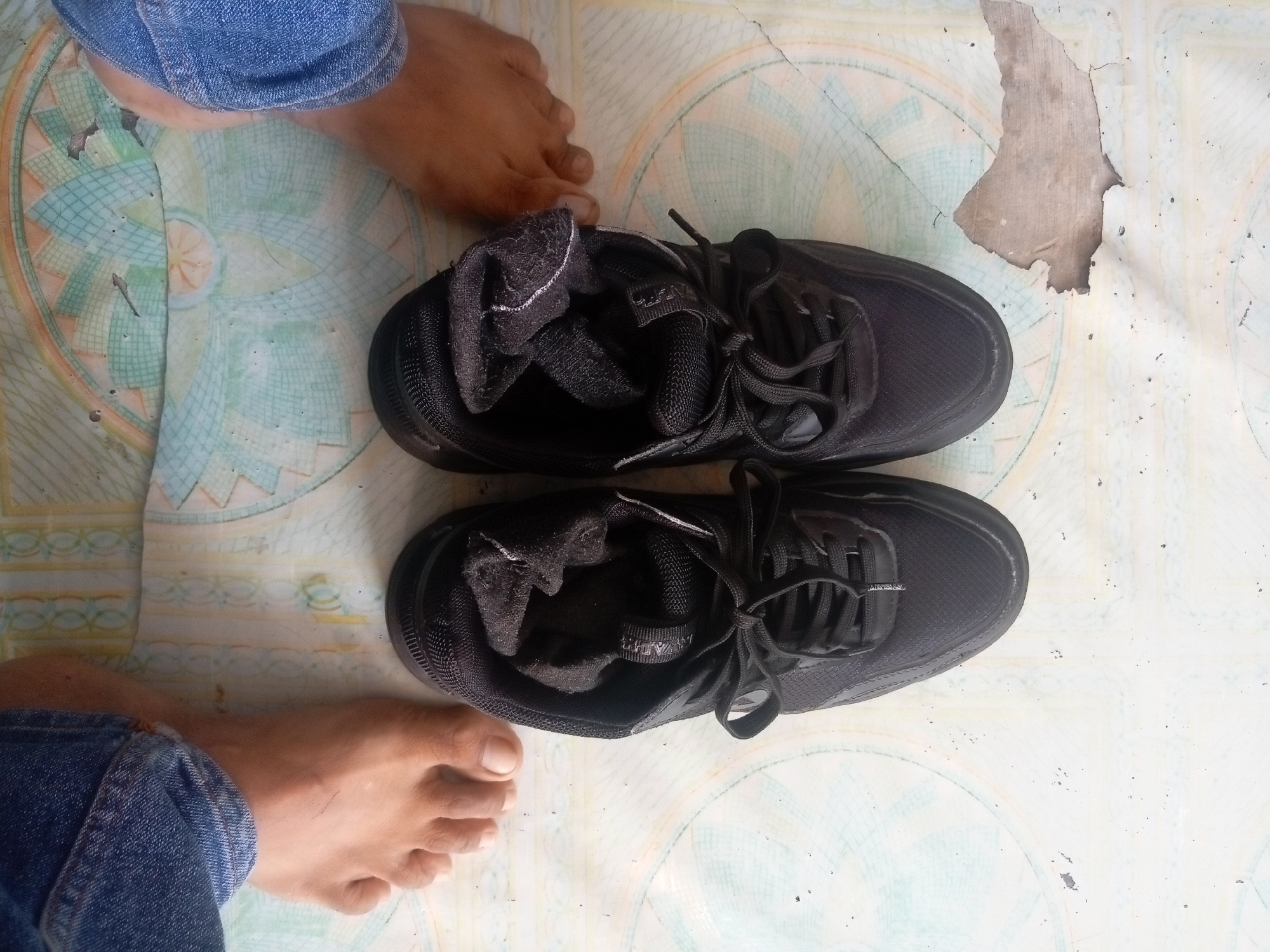 The image shows a pair of black athletic shoes placed on a patterned floor. The shoes are positioned horizontally in the center of the frame, with the toes pointing to the right. One shoe is slightly more open, revealing a black sock with some white trim inside. Two bare feet are visible, one at the top left of the image and another at the bottom left. The foot at the top is partially covered by blue denim jeans. The floor appears to be linoleum or a similar material with a repeating circular and diamond pattern in shades of green, beige, and white. There is a section of the floor near the top right corner that is damaged and peeling. The lighting suggests an indoor environment. No specific location in Batam, Indonesia, can be identified from the image.