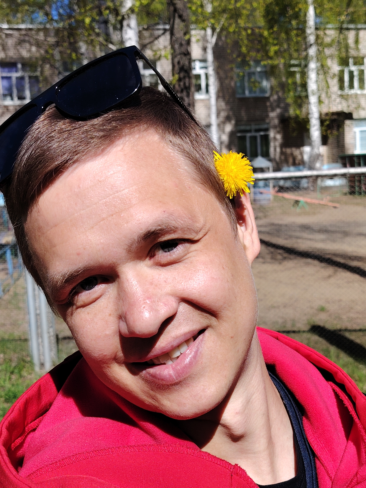 A close-up shot reveals a smiling man outdoors in Chaikovsky, Russia. Sunglasses are perched on top of his head, and a yellow dandelion is tucked behind his ear. He wears a red hooded sweatshirt over a black shirt. In the background, there's a building with multiple windows and tree branches. A playground, including what appears to be a seesaw, is visible in the blurred distance. A metal fence is also partially visible in the foreground. The scene is bathed in bright sunlight, creating shadows on the man's face and the surrounding environment.