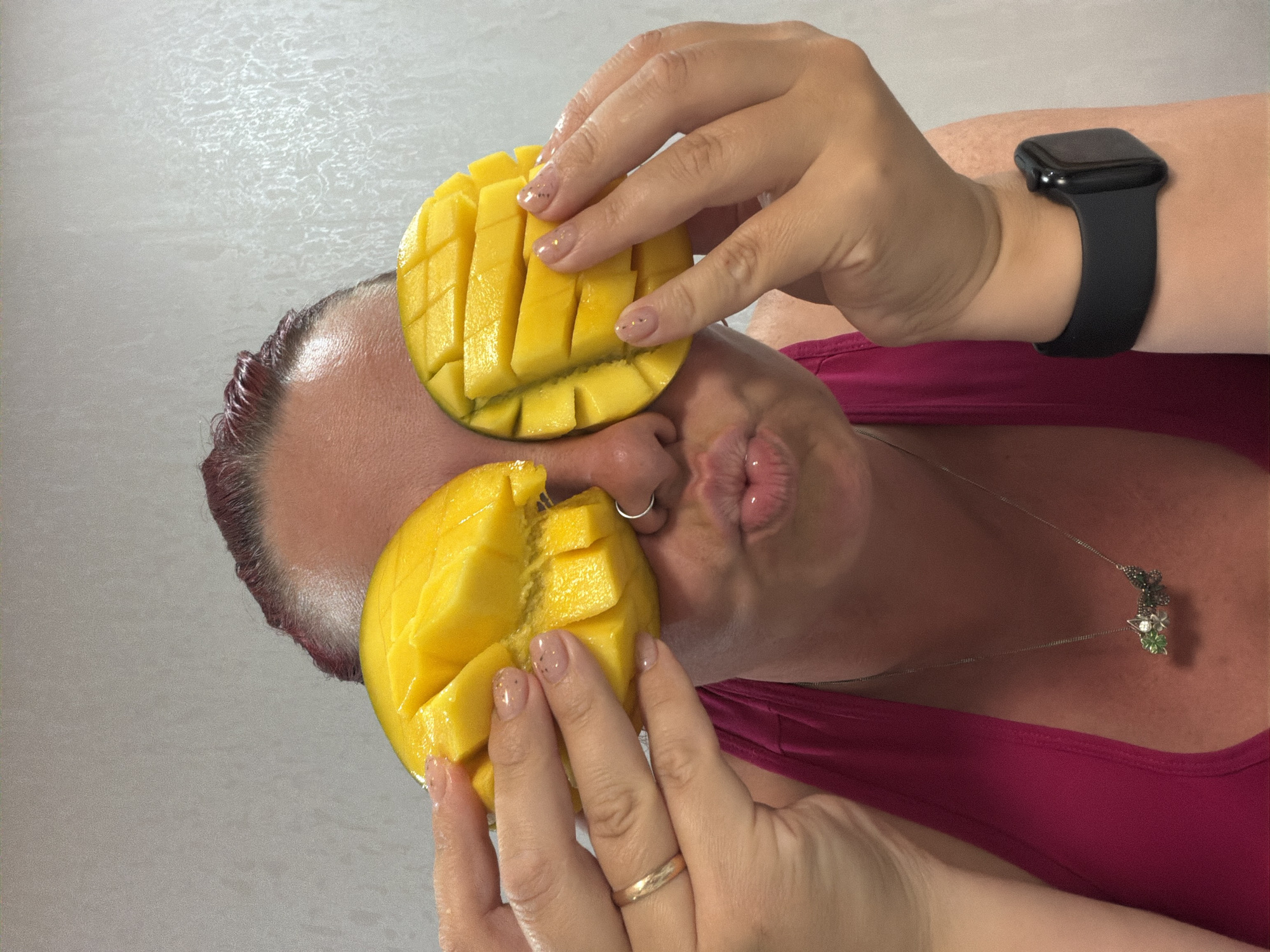A woman wearing a magenta tank top holds two cut mango halves, one over her eye and the other near her mouth. She has a nose ring and wears a necklace with a clover-shaped pendant. A smart watch is visible on her wrist. The background is a plain, light surface. The location is not discernible from the image.