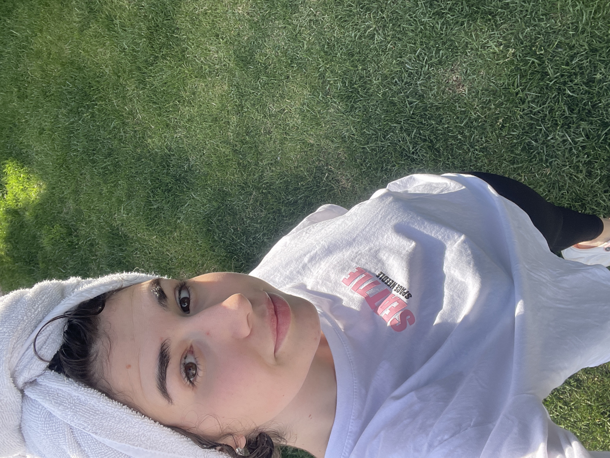 A young woman is lying on a grassy surface, likely a lawn. She is wearing a white t-shirt with the words "Seattle Space Needle" printed on it. A white towel is wrapped around her head. She is also wearing black leggings. The setting appears to be outdoors in daylight. The location in Sofia, Bulgaria, cannot be determined from the image.