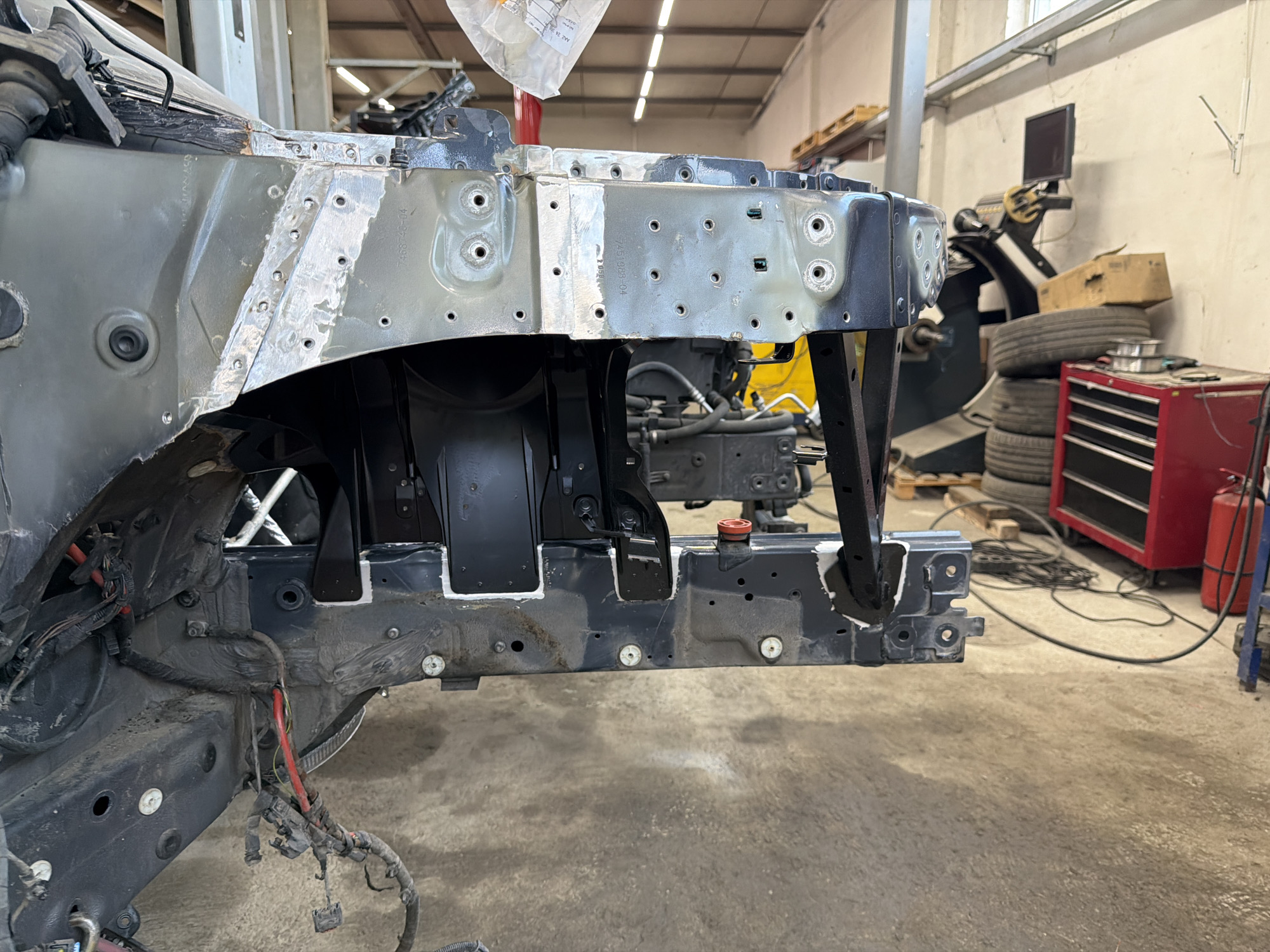 The image depicts the front end of a vehicle undergoing repair in a garage setting. The car's front structure has been partially disassembled, revealing metal framework, various holes, and remnants of wiring. Patches of light gray or silver indicate possible welding or repair work. In the background, tires are stacked next to a red toolbox, a tire balancing machine and other garage equipment. The setting appears to be a well-lit auto repair shop.