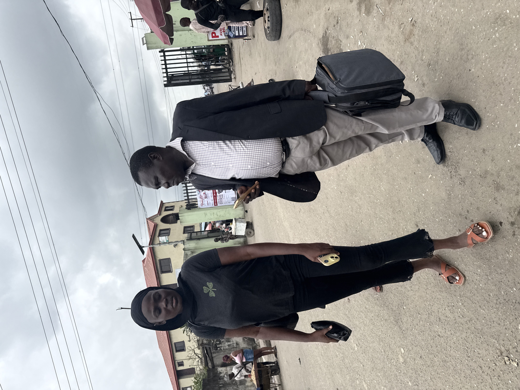 In Ado, Nigeria, two individuals, a woman and a man, are captured in a medium shot on a paved street. The woman, on the left, wears a black hijab and a black t-shirt adorned with a green shamrock emblem. She is also wearing black pants and brown open-toed sandals. She is holding a black clutch in her left hand and a yellow smartphone in her right. The man, on the right, is dressed in a dark suit jacket over a checkered dress shirt and light grey trousers. He is wearing black dress shoes and carrying a dark grey messenger bag in his left hand. He is holding a brown object, possibly a pen or stylus, in his right hand and appears to be looking towards his left. In the background, a gated entrance to a building complex is visible, along with a red-awninged stall and other individuals walking in the distance. The sky is overcast with visible power lines overhead.