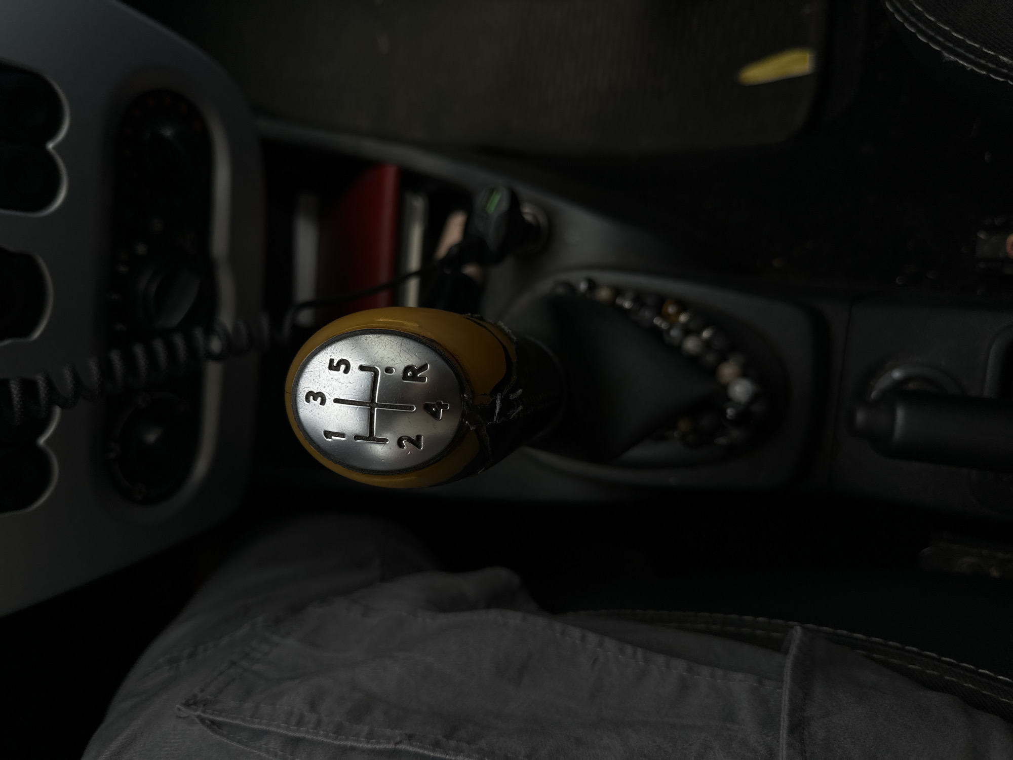 An overhead, close-up shot captures the interior of a manual transmission vehicle, focusing on the gear shift knob. The knob, a metallic silver disc with a yellow insert, displays the standard gear pattern: 1, 2, 3, 4, 5, and R. The lever is positioned in the neutral gear. To the left of the gear shift, a person's gray pants are visible, indicating the presence of a driver or passenger. In the background, dark car interior elements such as the dashboard and center console are discernible. A string of dark-colored beads hangs from the gear shift. The lighting is dim, suggesting a low-light environment within the car. There is no indication of the specific city or location within Бараўлянскі сельскі Савет, Belarus.
