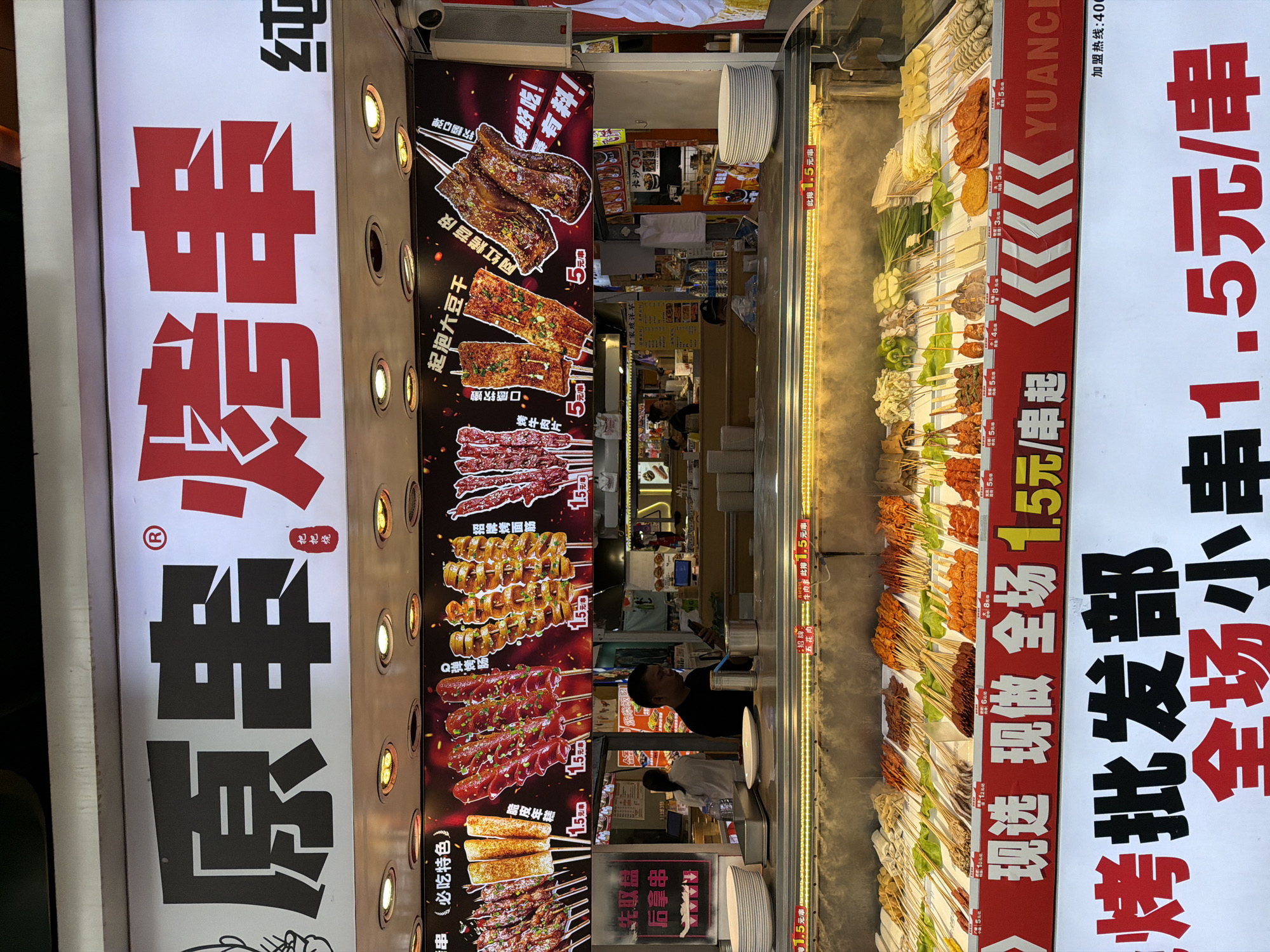 The image shows the storefront of a "Yuan Chuan" skewered meat stall, in the Chongqing High-tech Zone of China. The stall has two large signs on either side of a display of food items on skewers. The left sign has the name of the stall in large Chinese characters, "原串烤串". The signs in the middle show several prepared foods on skewers and their prices. The right sign advertises that "Whole market small skewer 1.5 yuan/skewer", and that it sells wholesale with a hotline for joining. There are two individuals working at the storefront. One is a male facing inside the store while on the phone, and the other is wearing an apron and facing down.