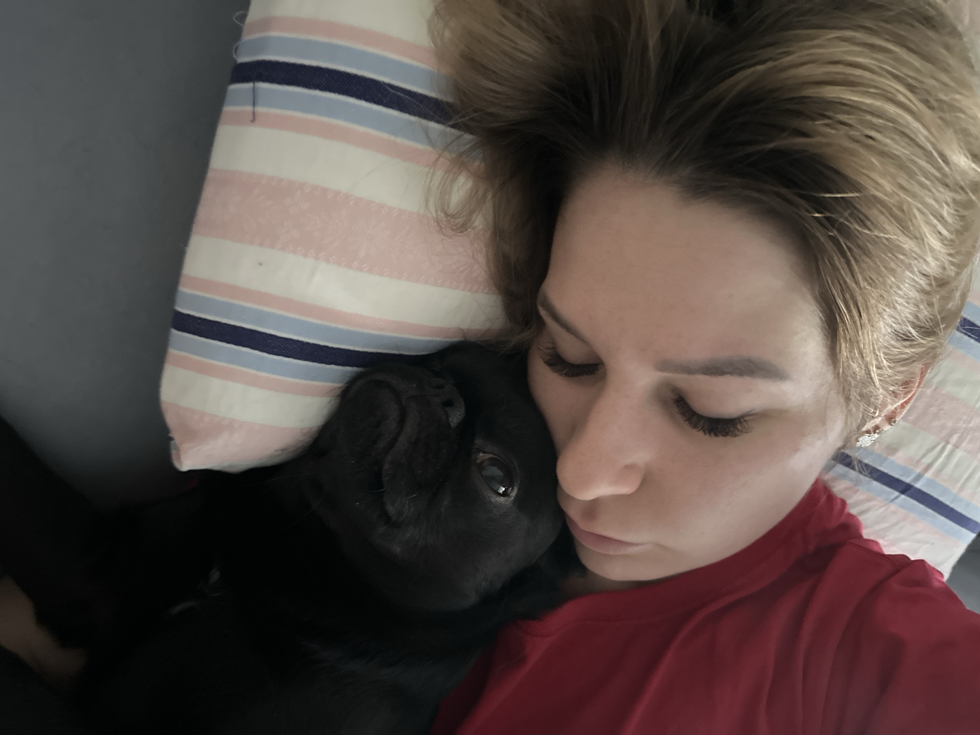 The image captures a close-up, overhead view of a person and a black pug dog snuggled together, likely in a bed. The person, appearing to be a woman, has light brown hair and fair skin. Her eyes are closed, suggesting she is sleeping or deeply resting. She is wearing a red t-shirt and has a small, decorative earring in her visible right ear. Her face and part of her hair are resting on a pillow adorned with white, light pink, light blue, and dark blue stripes.
Adjacent to the person's head and neck is a black pug, its head nestled against her. The pug's fur is dark, and one of its eyes is open, looking directly towards the viewer, adding a touch of alertness to the otherwise serene scene. The background on the left side of the frame appears to be a plain grey surface, likely a bed sheet or blanket. The lighting is soft and ambient, consistent with an indoor setting, possibly morning or evening. The overall impression is one of comfort, companionship, and peaceful co-existence between the person and their pet. There is no visible text in the image.