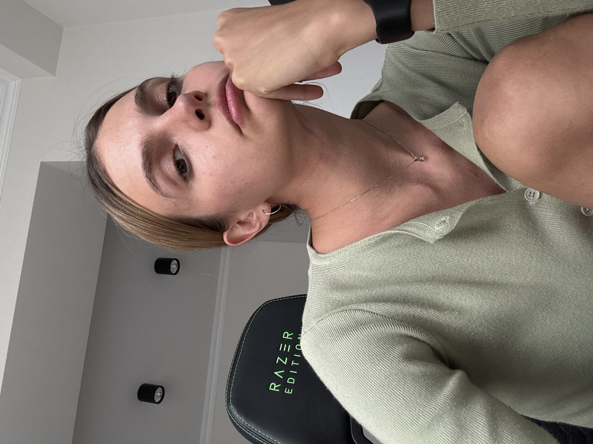 A young woman is captured in a medium close-up, looking directly towards the viewer with a thoughtful expression. Her head is slightly tilted, and her right hand rests near her chin or lower lip in a pensive gesture. She has light brown or blonde hair, styled away from her face, and her fair skin shows minimal makeup. She is wearing a light sage green ribbed cardigan or long-sleeved top, a delicate silver chain necklace with a small pendant, a small silver hoop earring in her left ear, and a black wristband on her right wrist.
The background consists of a plain, light-colored wall (likely white or light gray), against which two modern black cylindrical wall-mounted lights are visible. Part of a black gaming chair with green accents is visible to the lower left, bearing the text "RAZER EDITION" vertically in green capital letters. The overall setting appears to be indoors, possibly a modern room or home office. The lighting is soft and even, highlighting her features. Part of her upper arm and knee are also visible, suggesting she might be seated.